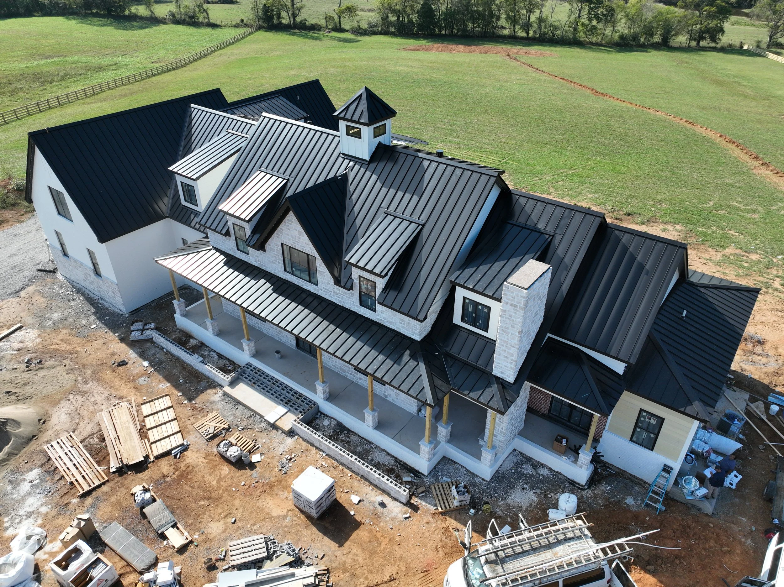 Aerial view of a house under construction with black metal roof, white exterior walls, and a small tower. Construction materials and tools are scattered around on dirt ground, and the house is surrounded by green fields and fencing.
