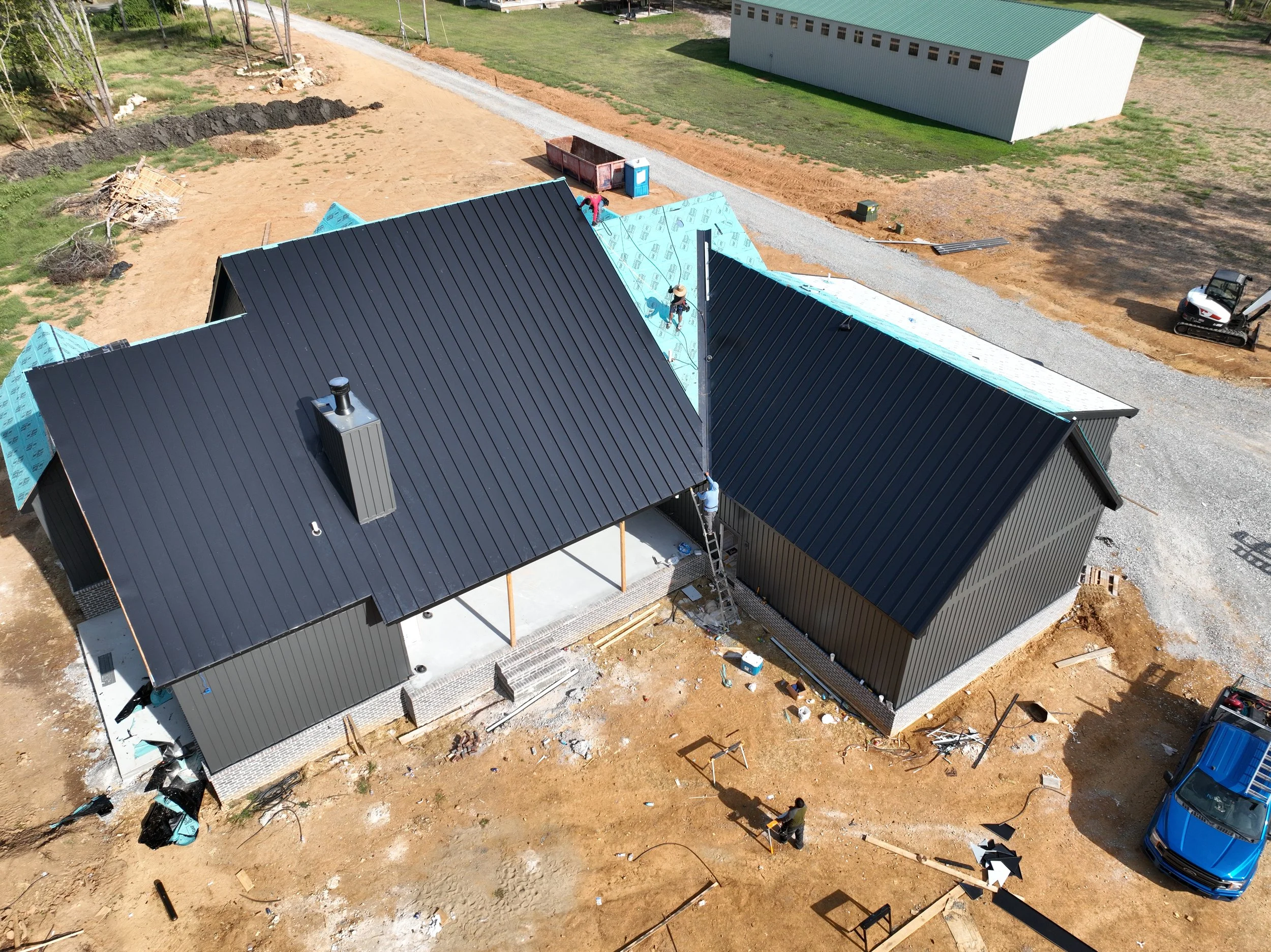Aerial view of a house under construction with a black metal roof, workers installing roofing, construction materials, and equipment around the site. A gravel pathway runs alongside the house, with a large white storage building in the background.