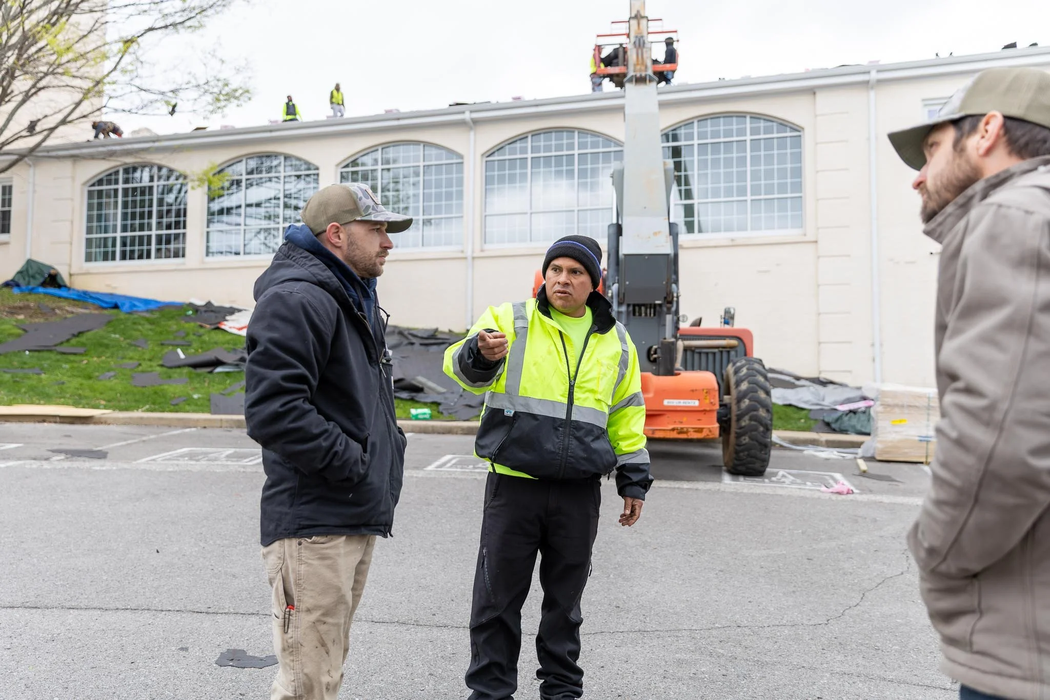 Three men are having a conversation outdoors near building repairs. One man is wearing a black jacket and beige pants, the second man is in a high-visibility yellow jacket and dark pants, and the third man, on the right, is in a beige jacket and cap. In the background, there are workers on a cherry picker working on the roof of a building, and construction materials are scattered on the ground.