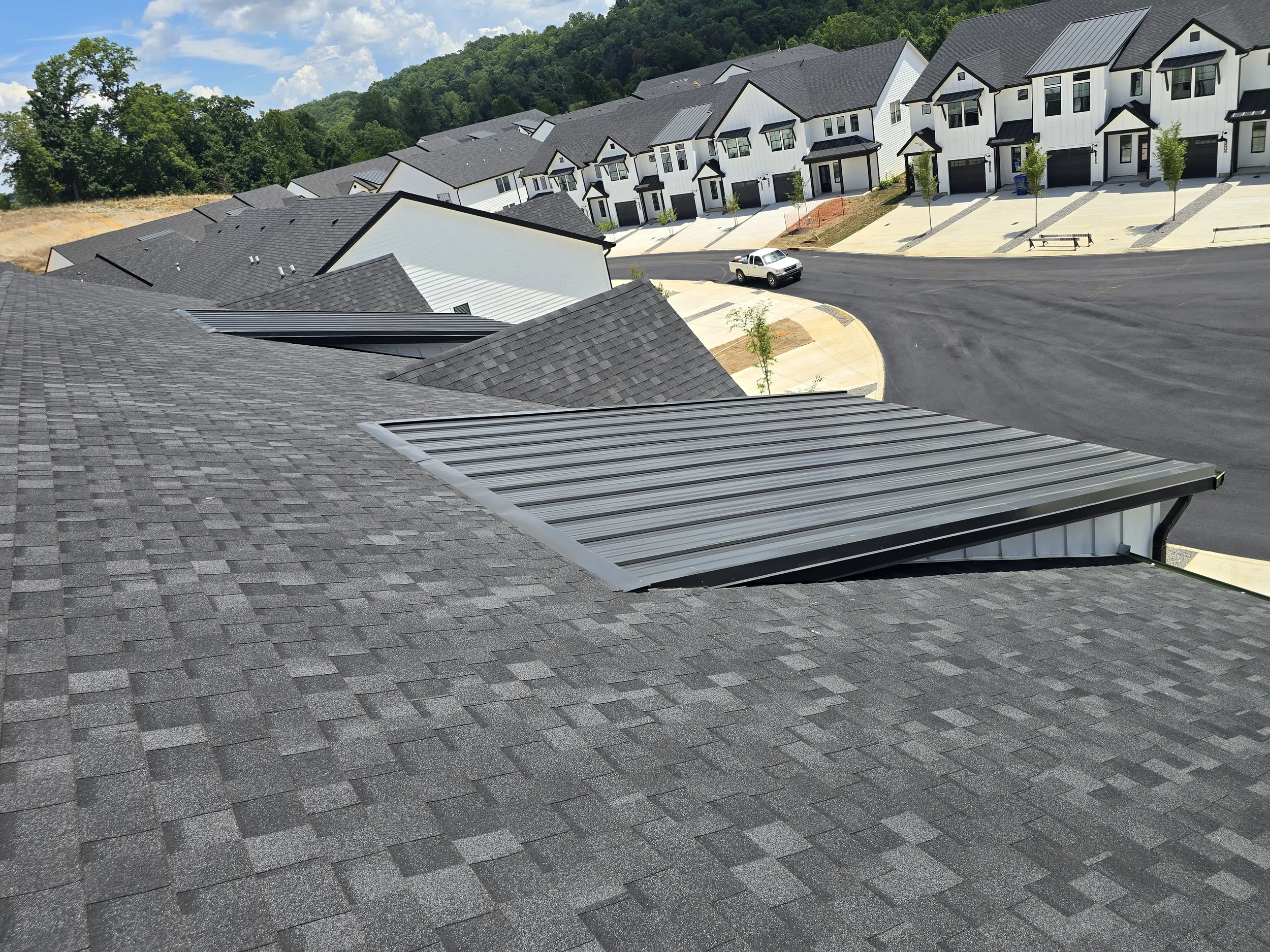 A row of modern white townhouses with black roofs and attached garages, situated along a curved asphalt street with newly planted small trees and a park bench. Hills and green trees are visible in the background under a partly cloudy sky.