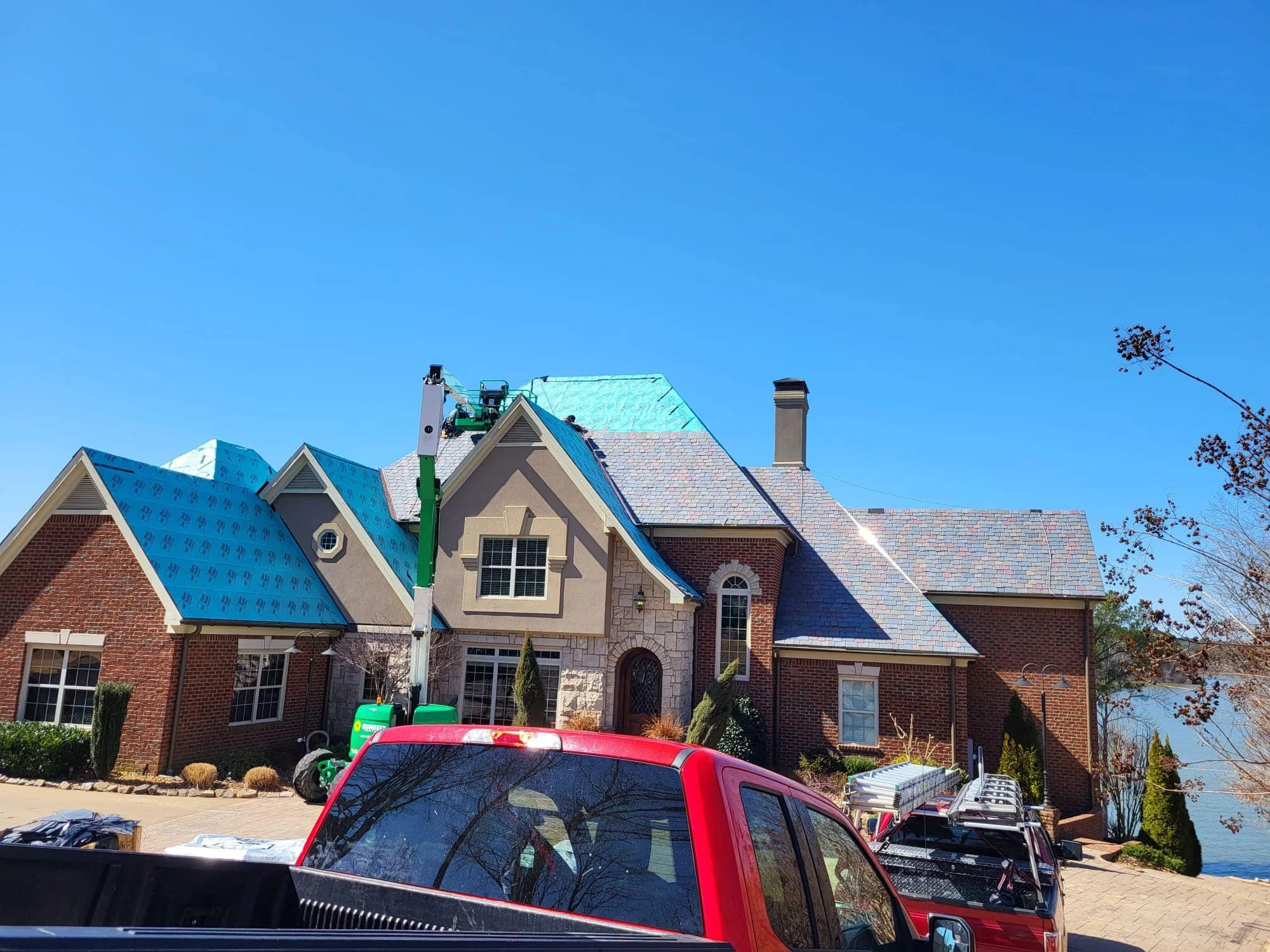 A house with a steep, brick roof is undergoing roofing work supported by a green lift. The house has a brick facade, multiple windows, and a stone section near the main door. There are parked cars in the driveway, and construction equipment visible, 