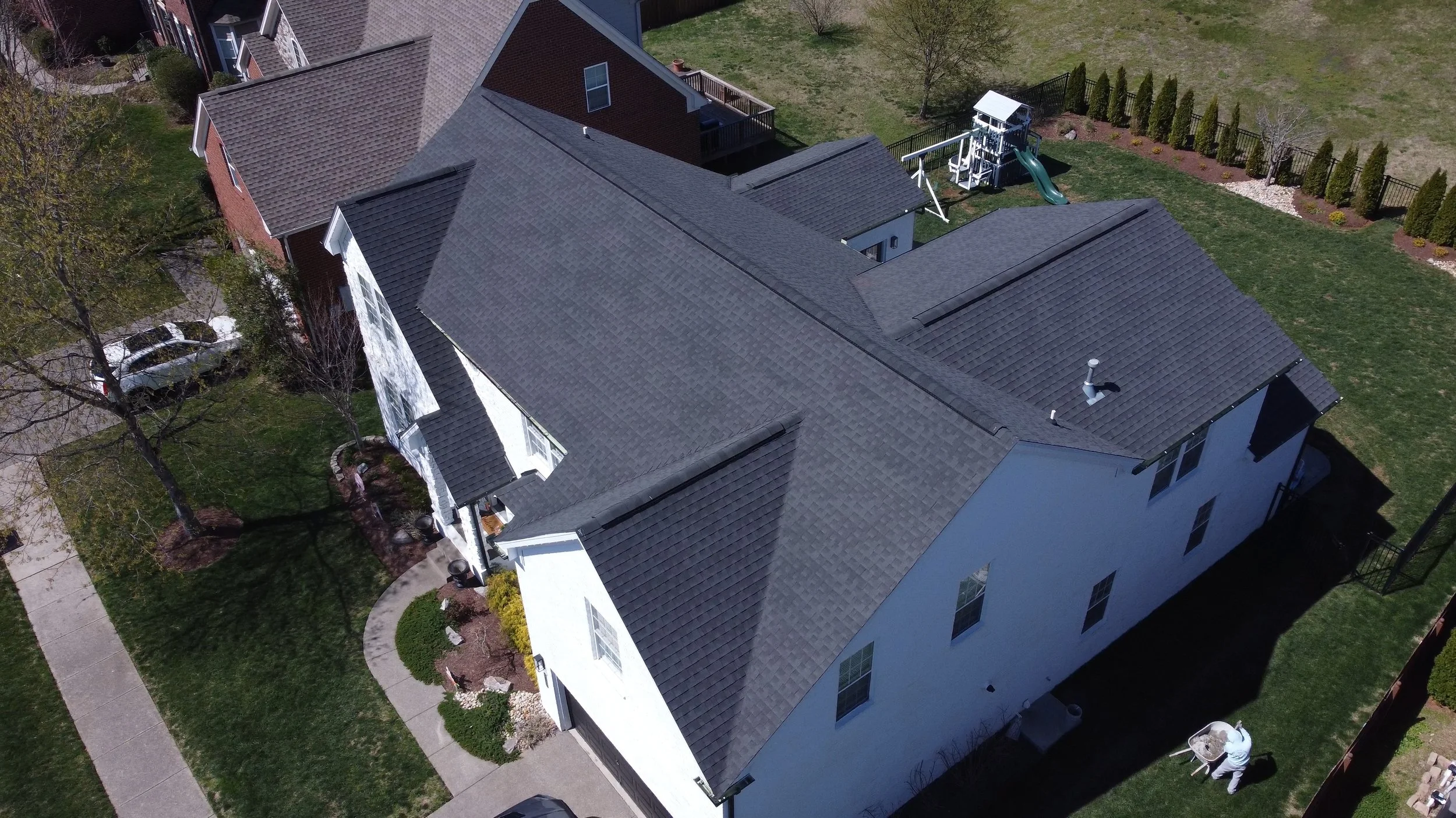 An aerial view of a two-story house with a dark gray roof, white exterior walls, and multiple windows. The house has a small front porch with stairs and a curved sidewalk leading to the street. The backyard features a playground with a slide, surroun