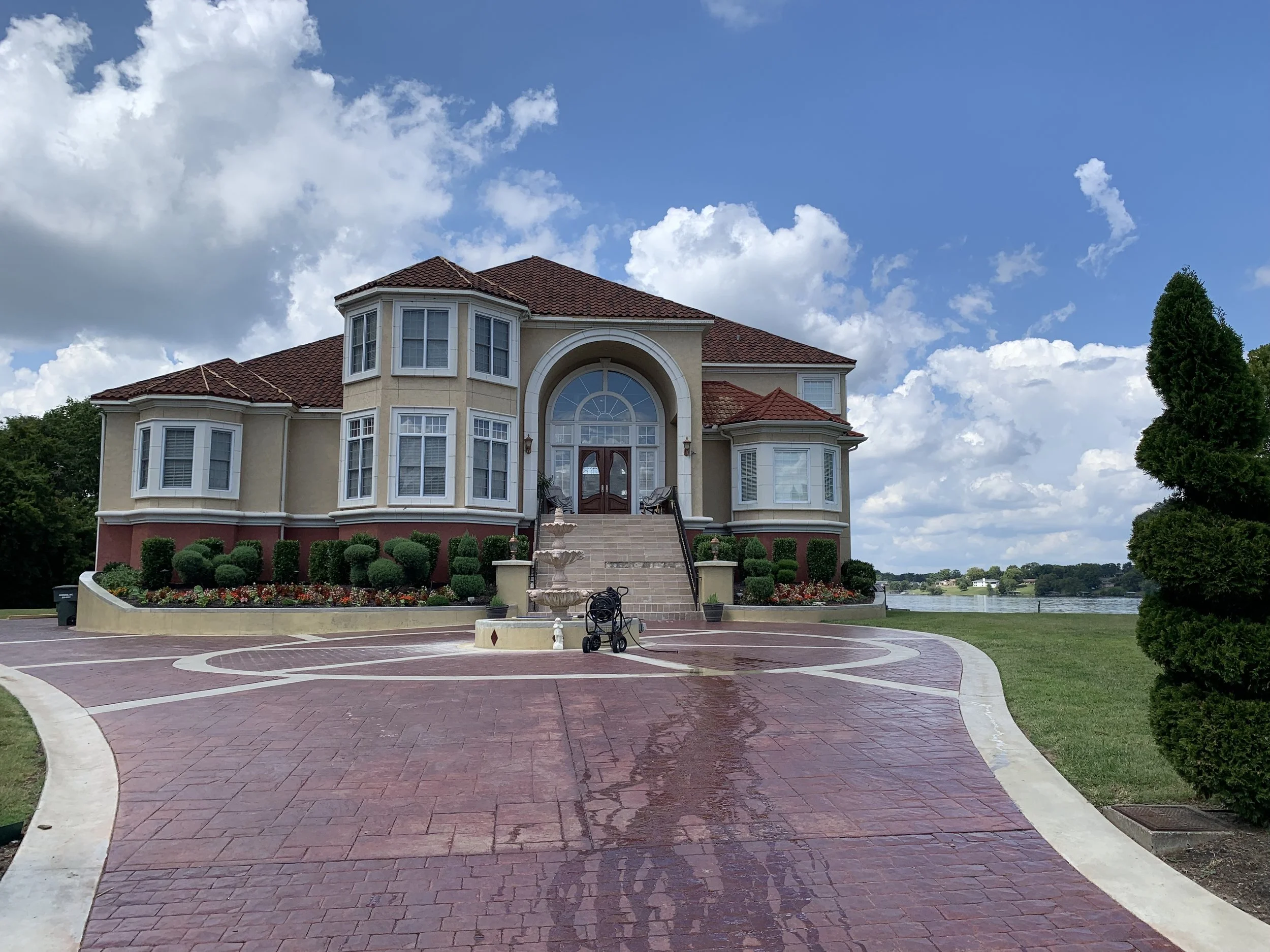 A large, beige house with a red tiled roof and multiple bay windows. There is a stairway leading up to a wooden front door with an arched window above. In front, a paved driveway with a fountain, landscaping, and a stroller. Trees and water body visi
