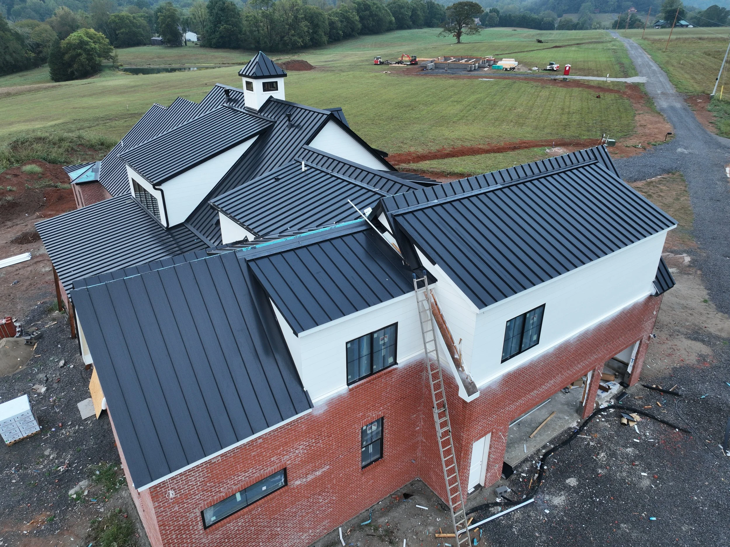 A black metal roof being installed on a white and red brick house in a rural area, with construction tools and equipment around.