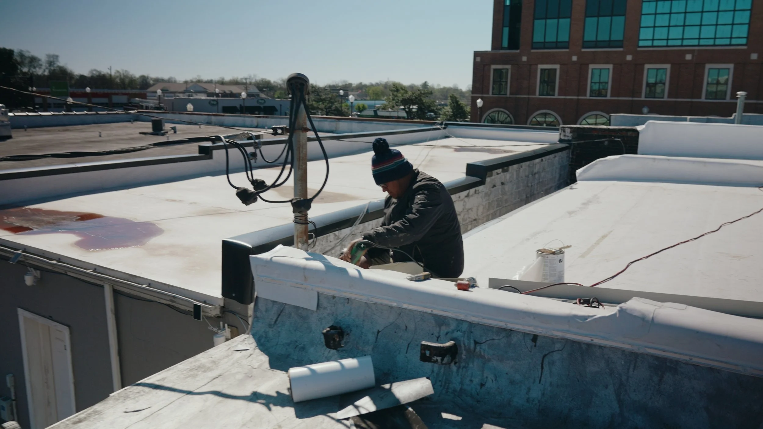Person wearing a striped beanie working on a flat rooftop with roofing materials and equipment, with buildings and trees in the background.