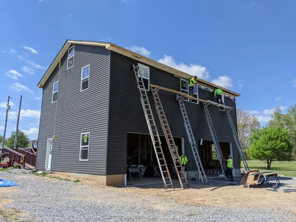Construction workers installing siding on the upper level of a black house with multiple ladders and scaffolding.