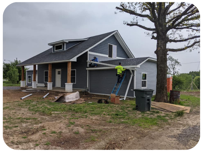 Construction workers installing siding on a house with a large tree in the yard.