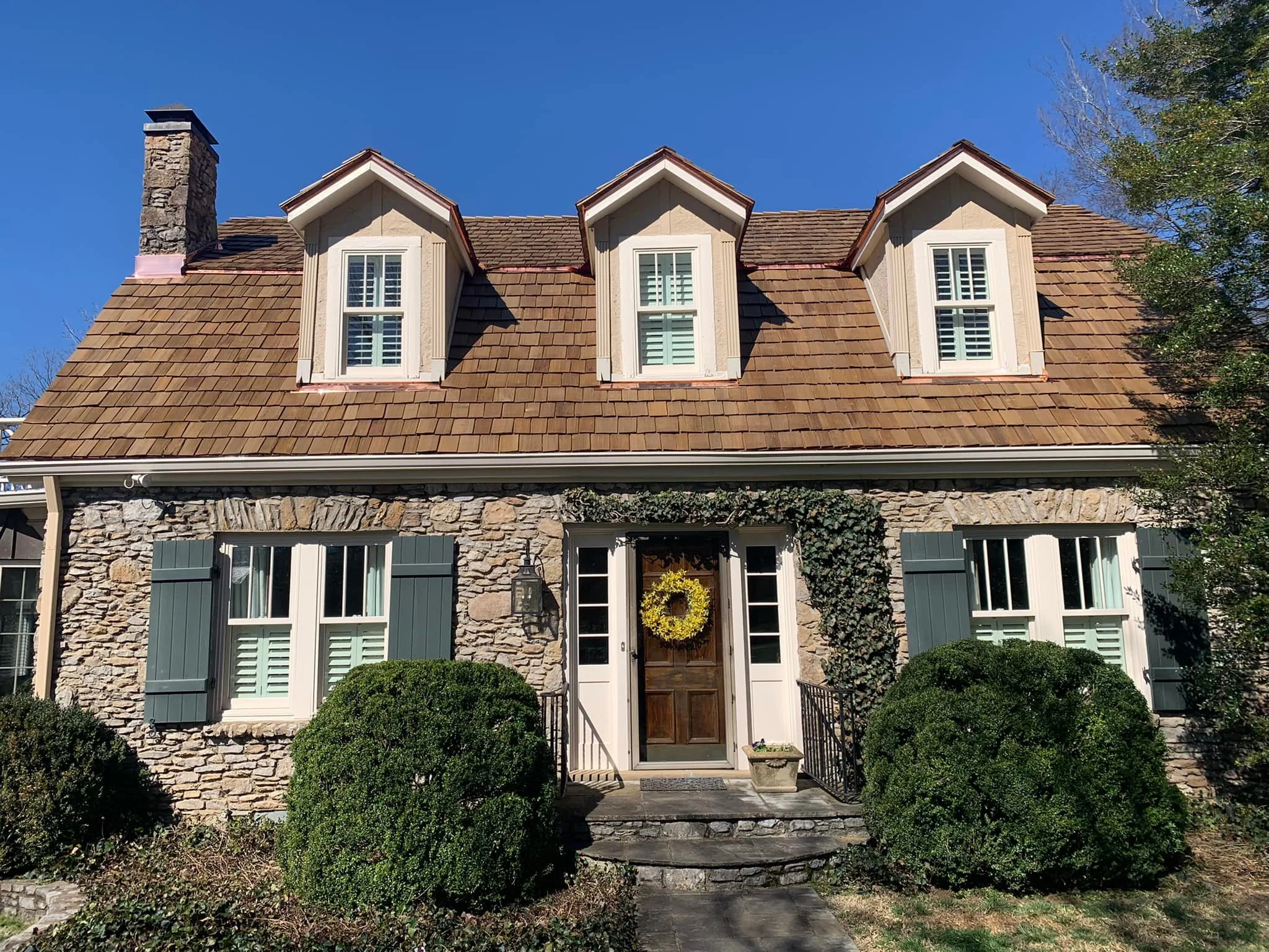 Front view of a house with a stone facade, three dormer windows, a wooden front door with a yellow wreath, shutters, and lush shrubs and bushes in the front yard.