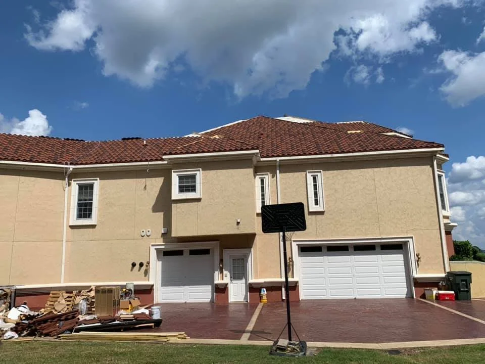 A two-story house with beige walls and a red tile roof, featuring multiple white-framed windows, a double garage, a basketball hoop in front, and a cloudy sky overhead.
