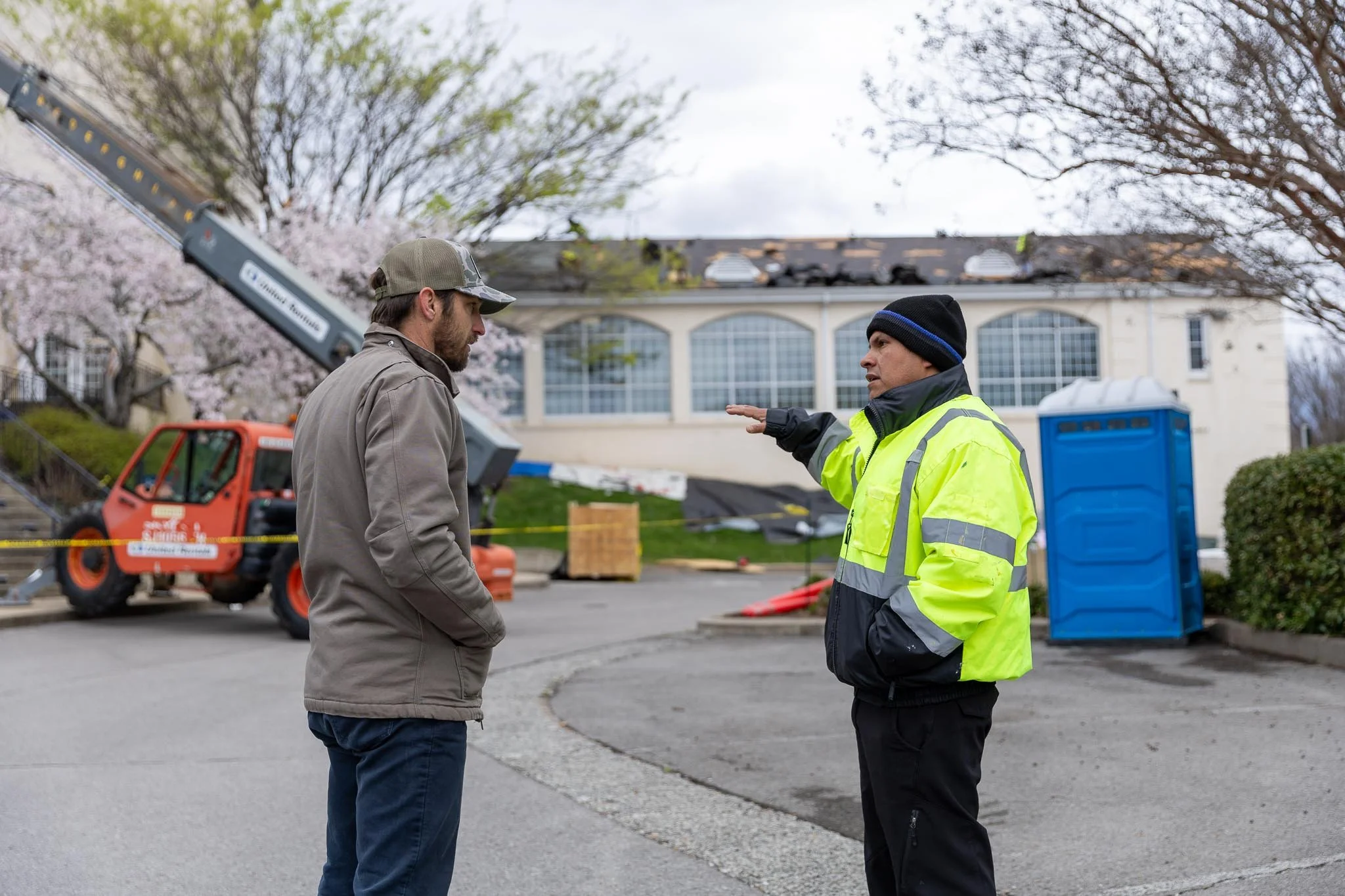 Two men conversing outdoors, one wearing a high-visibility yellow jacket and black gloves, the other in a tan jacket and baseball cap, with construction equipment and a building with large windows in the background.