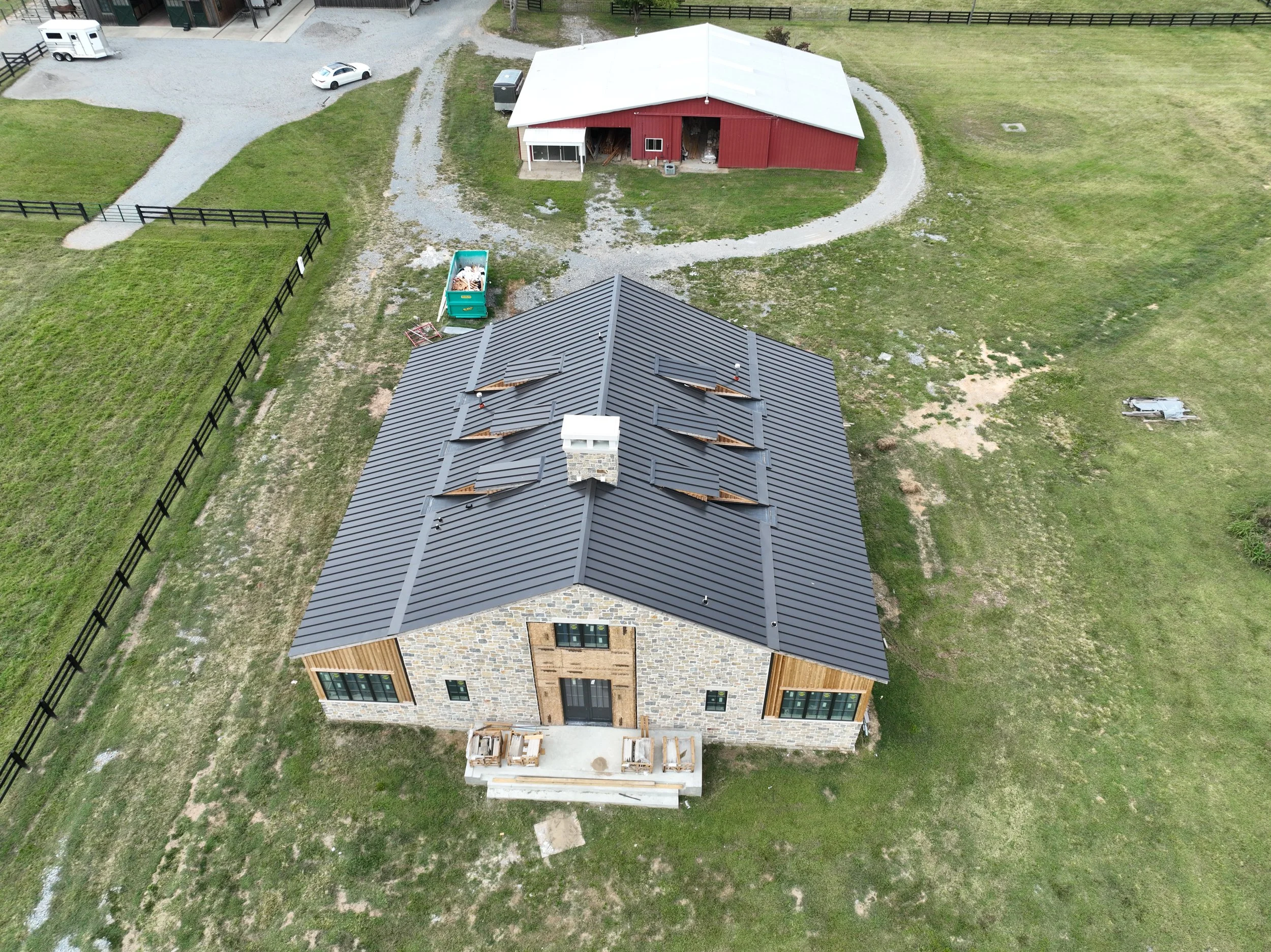 An aerial view of a two-story house with a gray metal roof, stone exterior, and a small front porch. The house is surrounded by green grass, a black wooden fence, and a gravel driveway. There is a trailer and some construction materials on the lawn. 