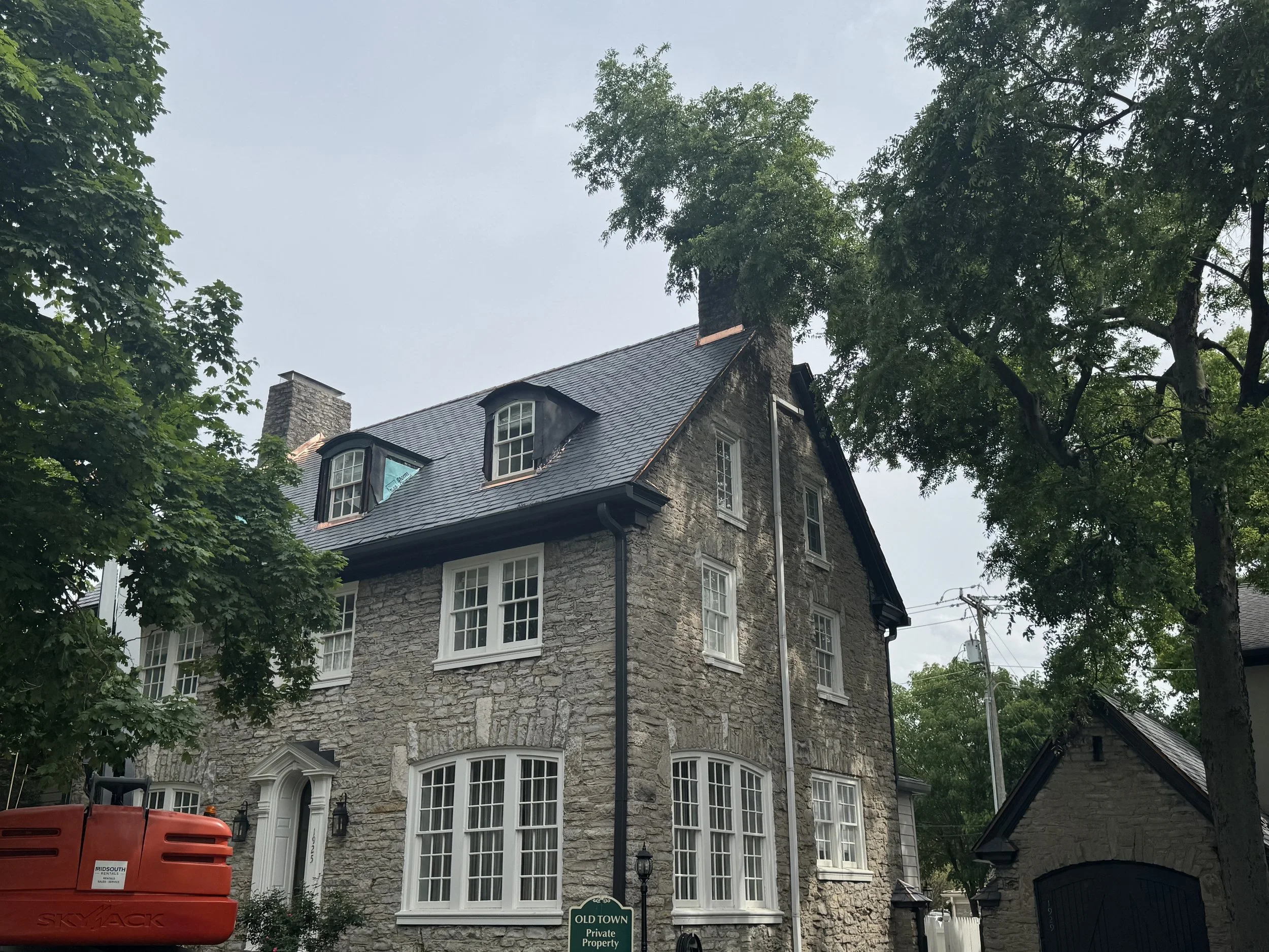 A large stone house with multiple windows, dormer windows on the roof, and a chimney, surrounded by tall trees.