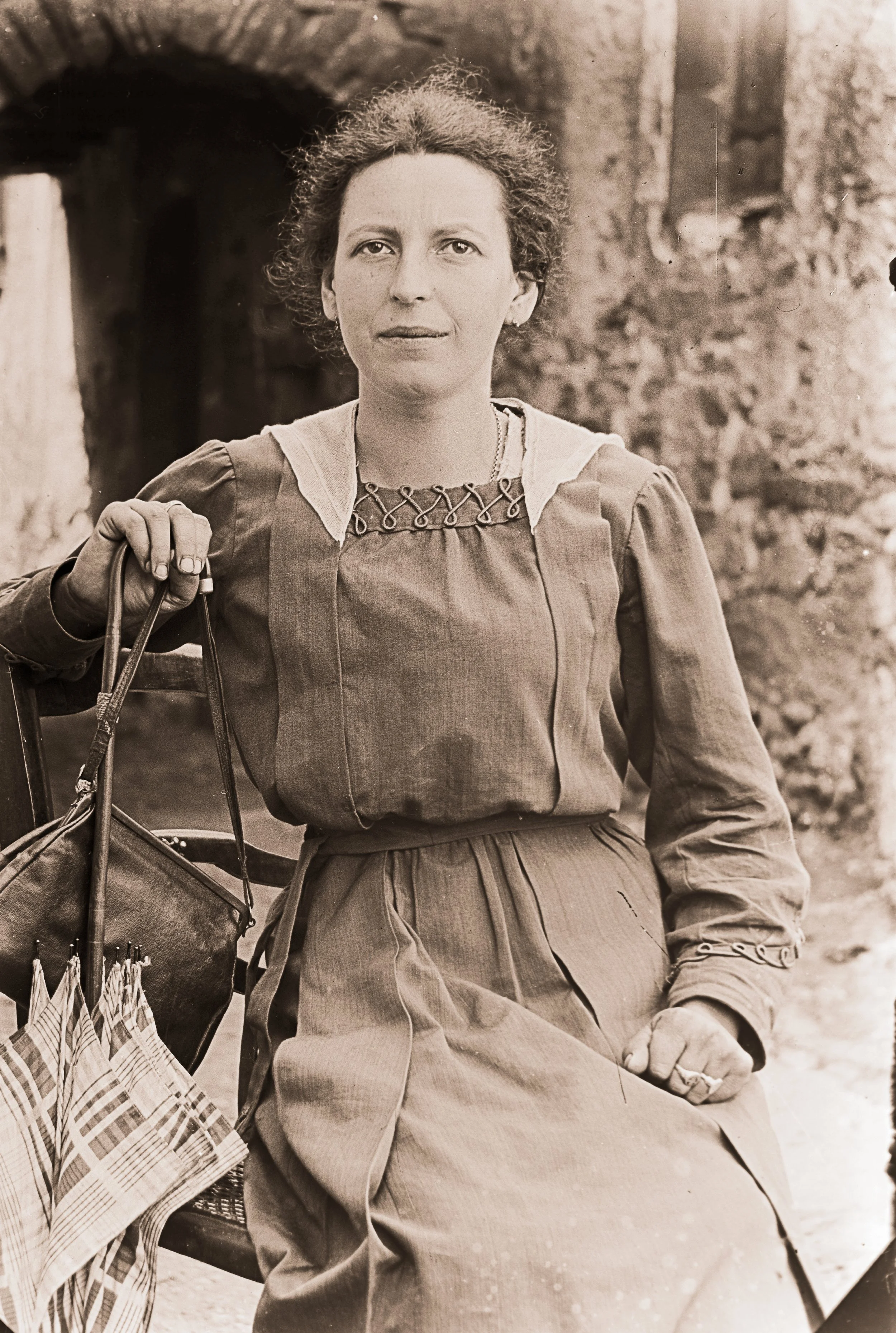 Femme assise avec un sac à main, portant une robe à manches longues et un collier, devant un mur en pierre.