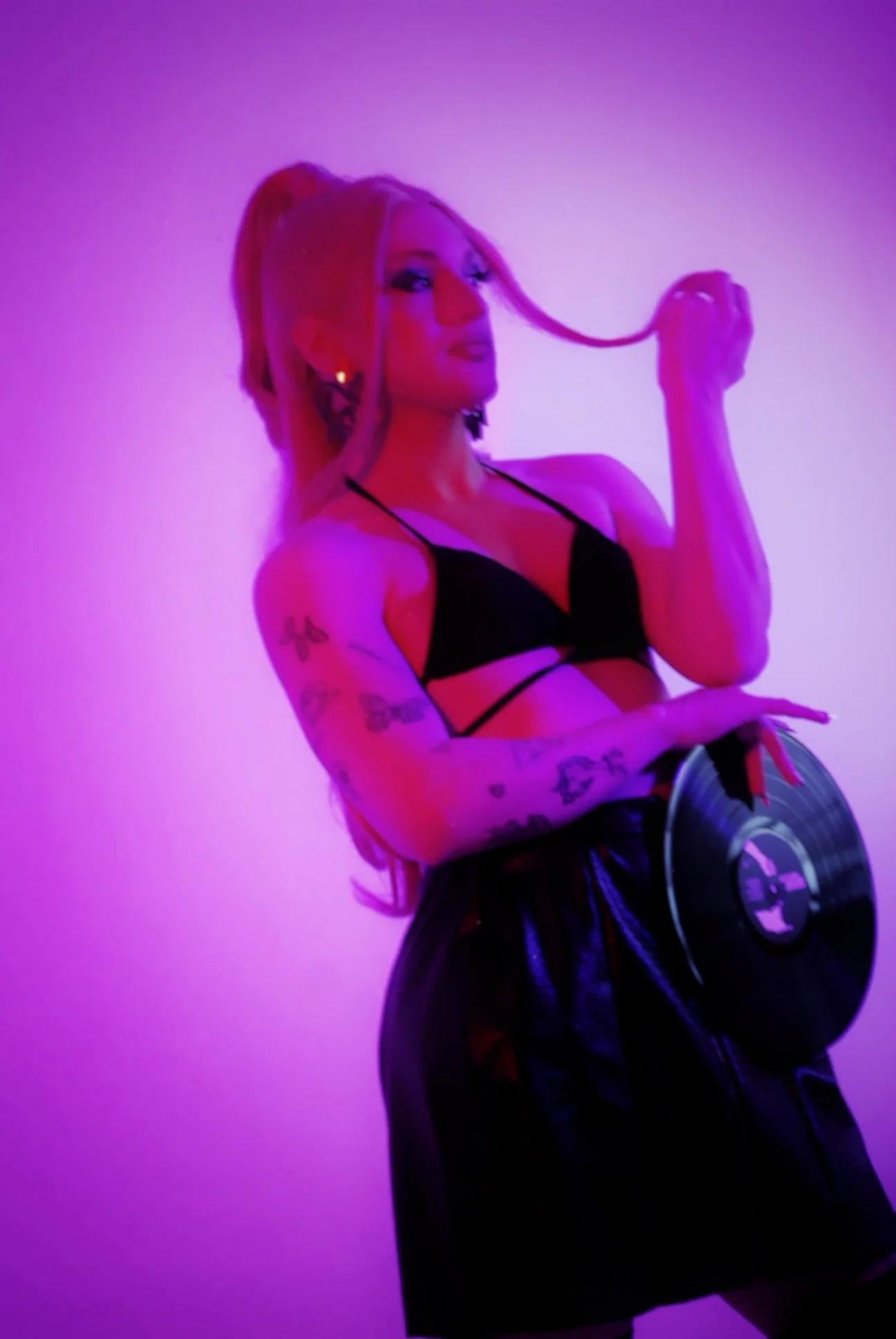 Mujer con tatuajes, vestido negro y con una cabina de discos en las manos, bajo luces moradas y rosadas.