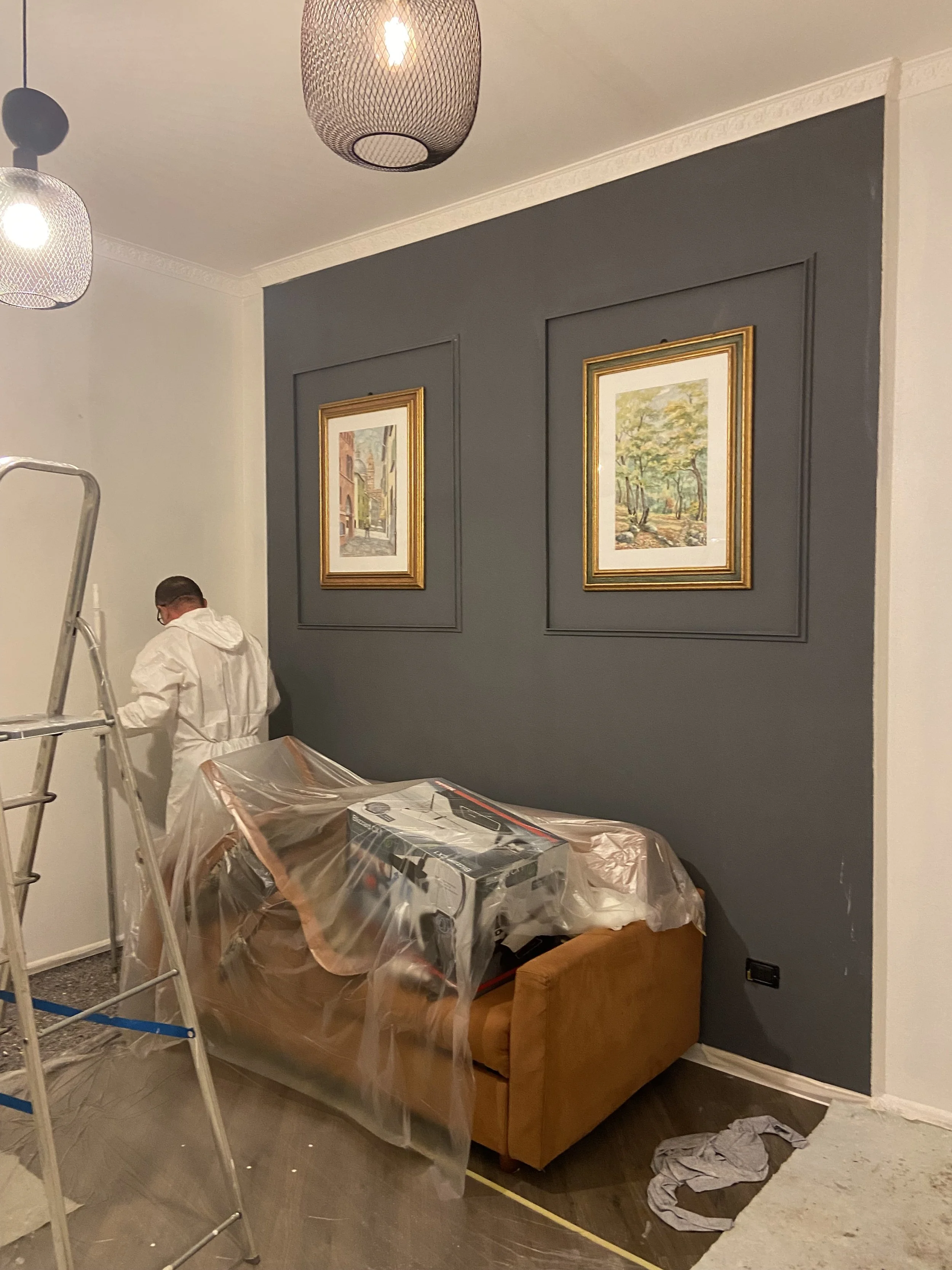 Una stanza in fase di ristrutturazione con un divano marrone coperto da plastica e un uomo in tuta bianca che lavora vicino a un muro grigio con due quadri con cornici dorate.