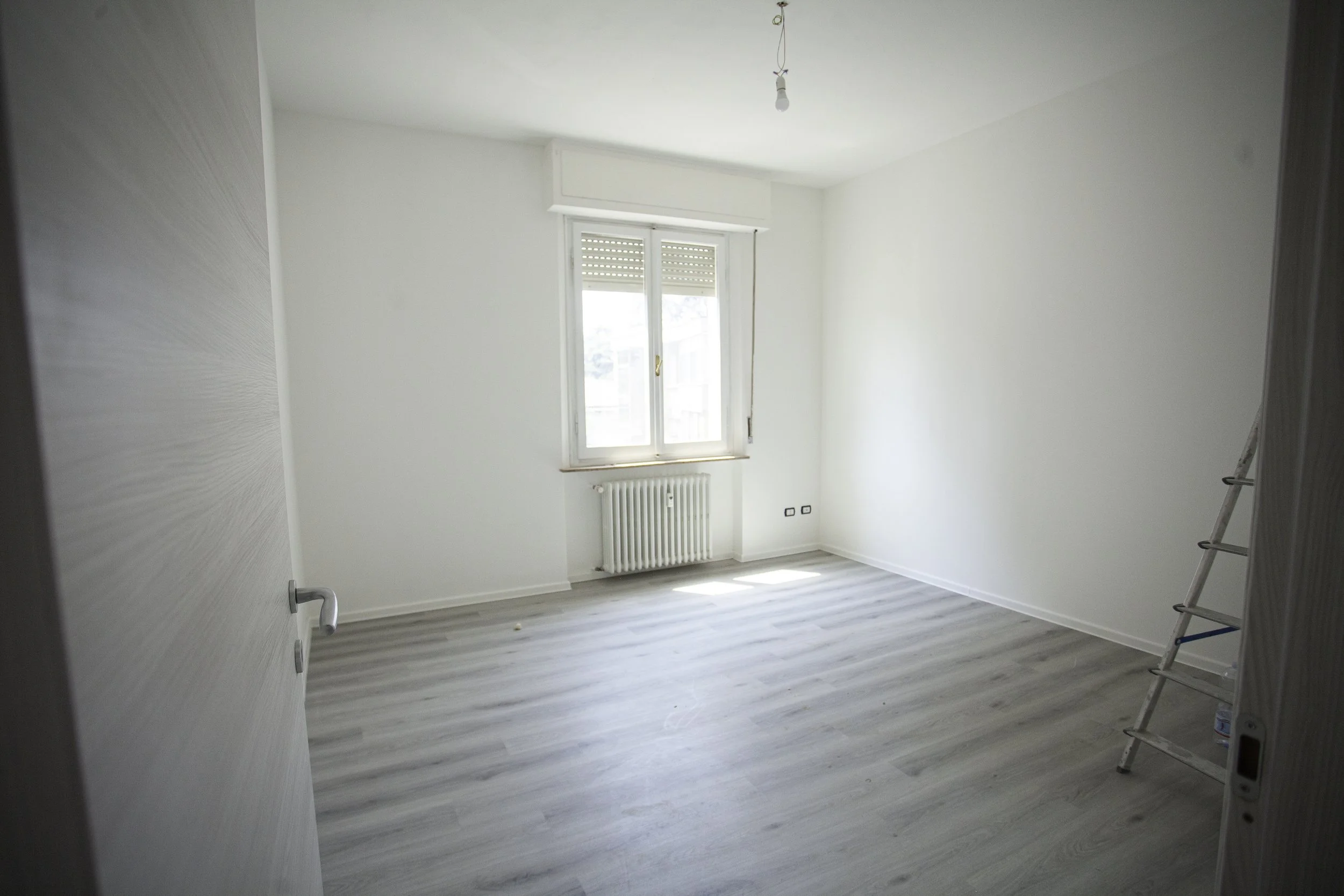 Stanza vuota con pareti bianche, finestra grande, termosifone sotto la finestra, pavimento in laminato chiaro, una scala stando di fianco alla parete destra.