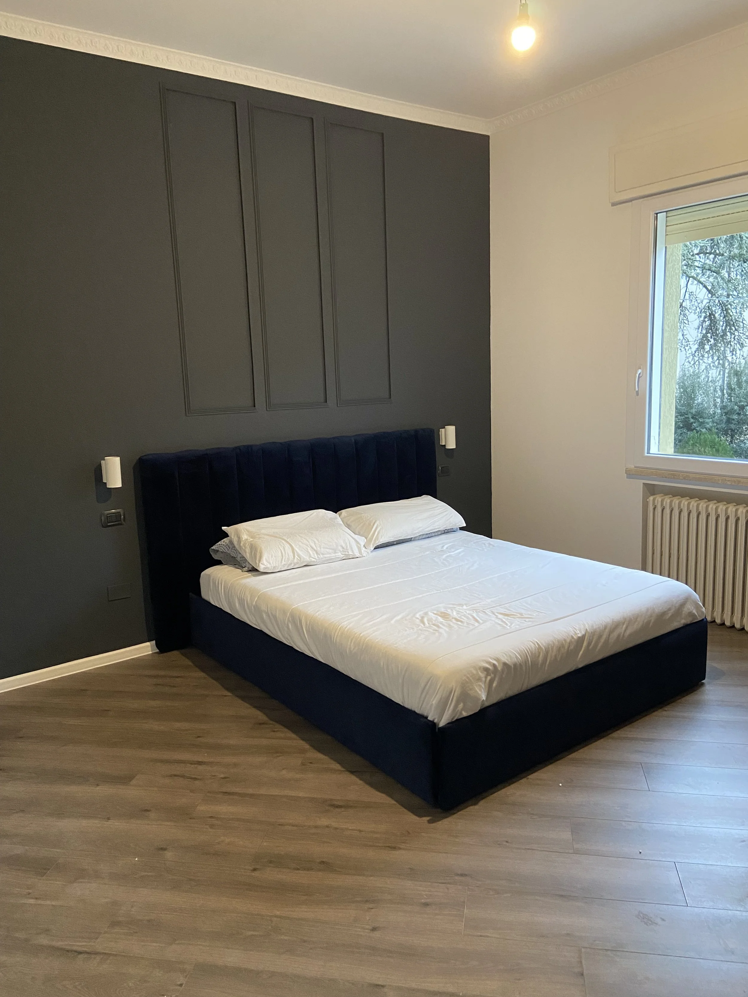 Una camera da letto con un letto con una testiera imbottita di colore blu, lenzuola bianche e due cuscini. Sulla parete dietro il letto c'è una parete di colore grigio con alcuni pannelli decorativi, e vicino alla finestra, che mostra albero e natura esterna, c'è un radiatore.
