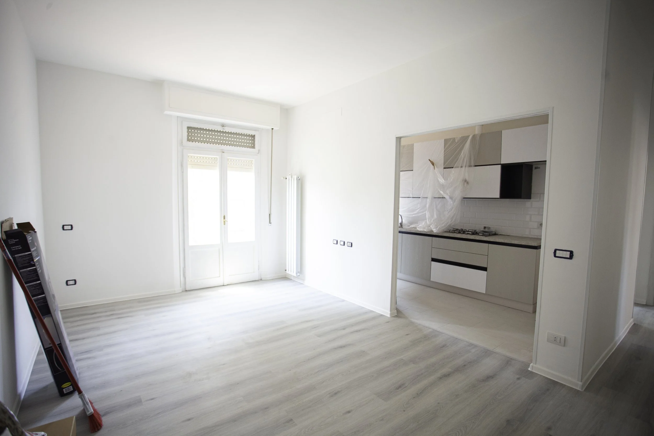 Interno di un appartamento in fase di ristrutturazione, con pareti bianche, pavimento in laminato chiaro, finestra con tapparelle, cucina moderna con mobili bianchi e pareti rivestite in piastrelle bianche, alcuni oggetti e strumenti di lavoro sul pavimento.