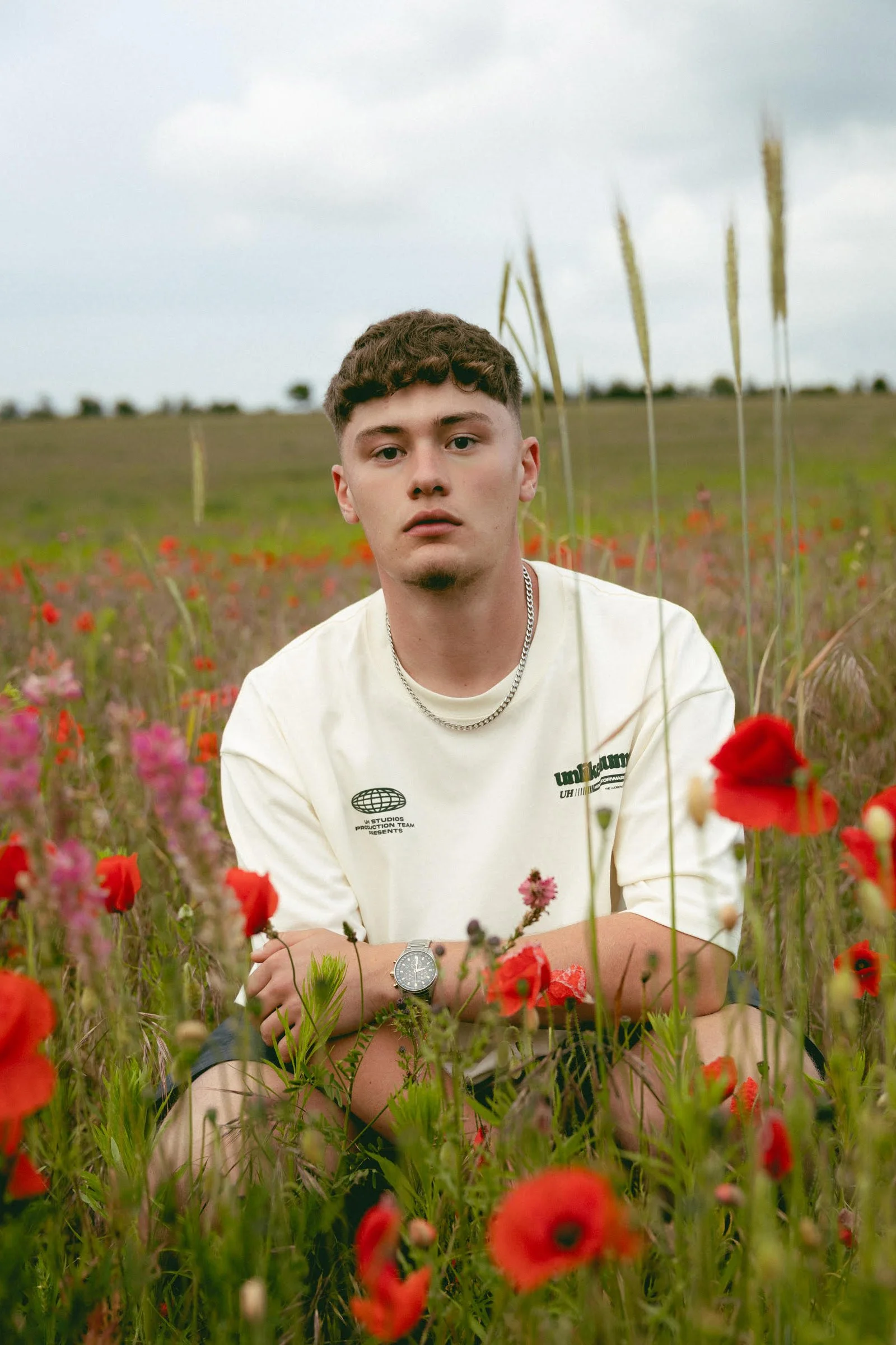 A young man with short brown curly hair and a goatee sitting in a field of wildflowers, including poppies and other colorful flowers, during daytime under a cloudy sky.