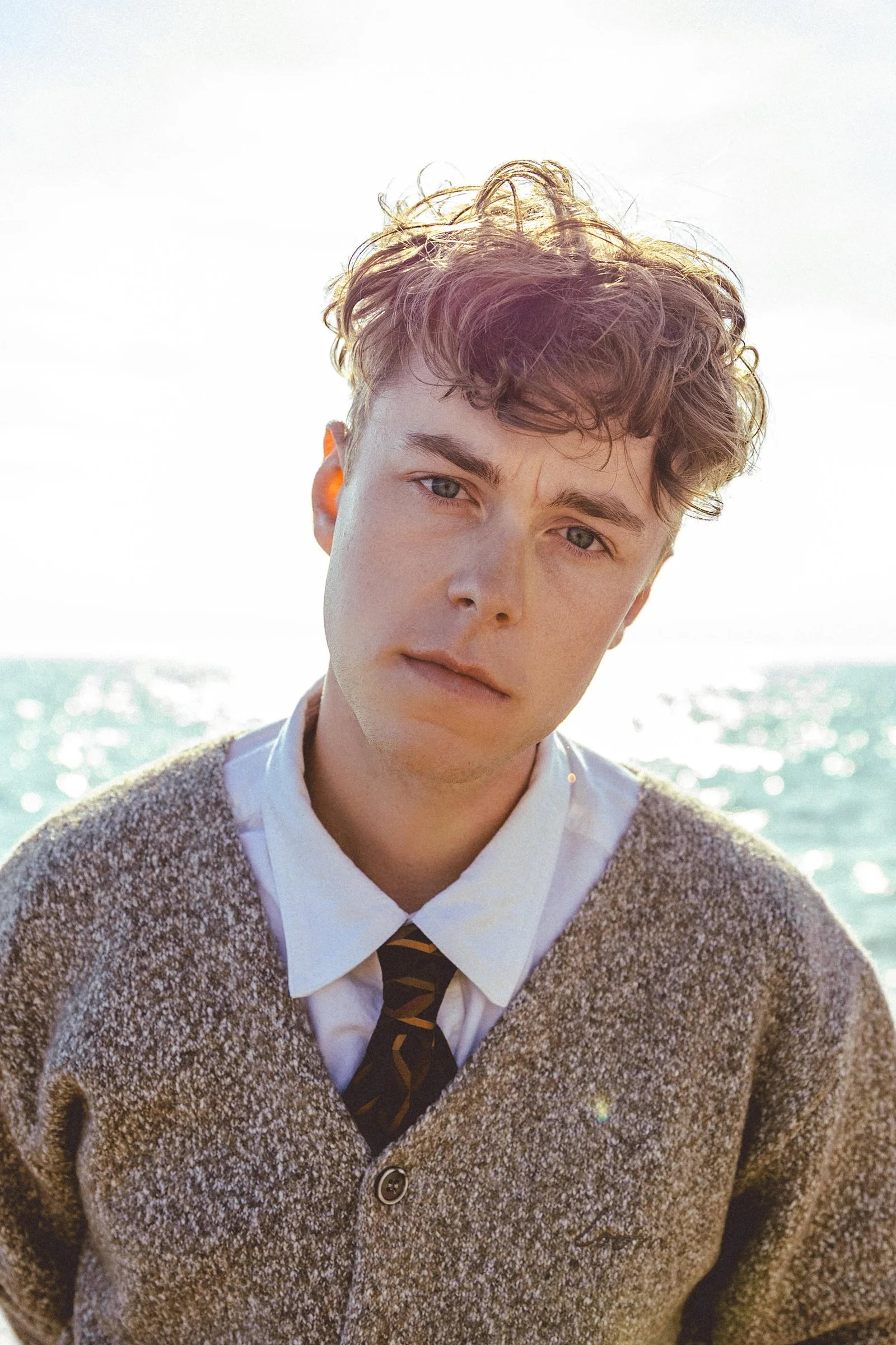 A young man with curly brown hair and light skin standing outdoors, dressed in a white collared shirt, a sweater, and a patterned tie, with a calm expression on his face. The background features a bright sky and sparkling water.
