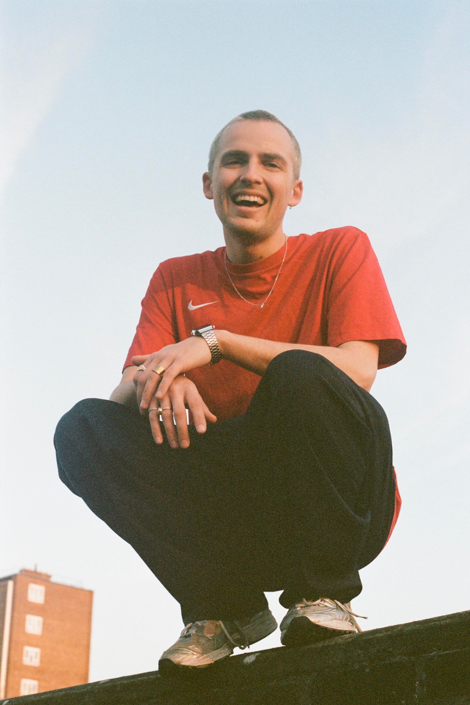Young man with short hair smiling while crouching on the edge of a rooftop with a building and sky in the background.