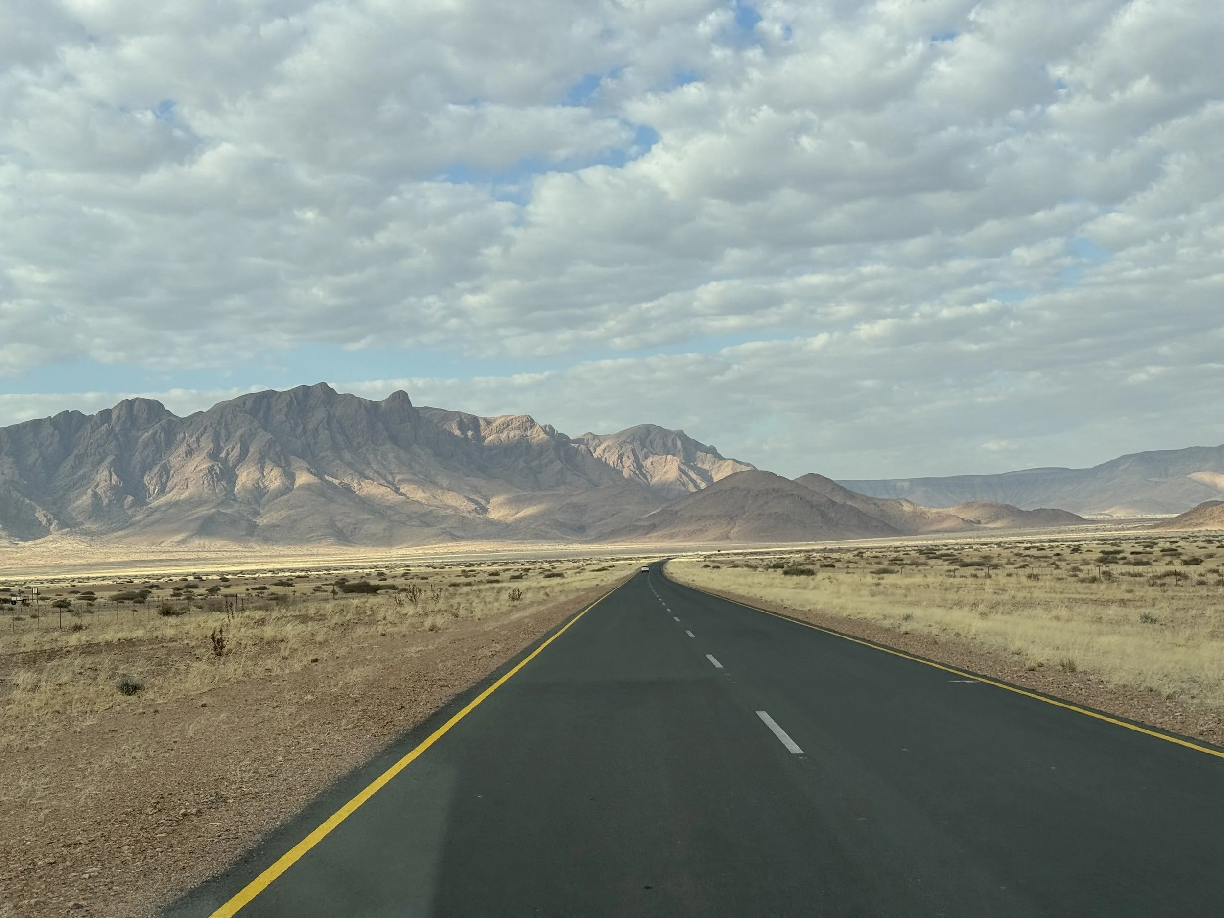 A long straight desert highway stretching towards mountains under a sky filled with scattered clouds.