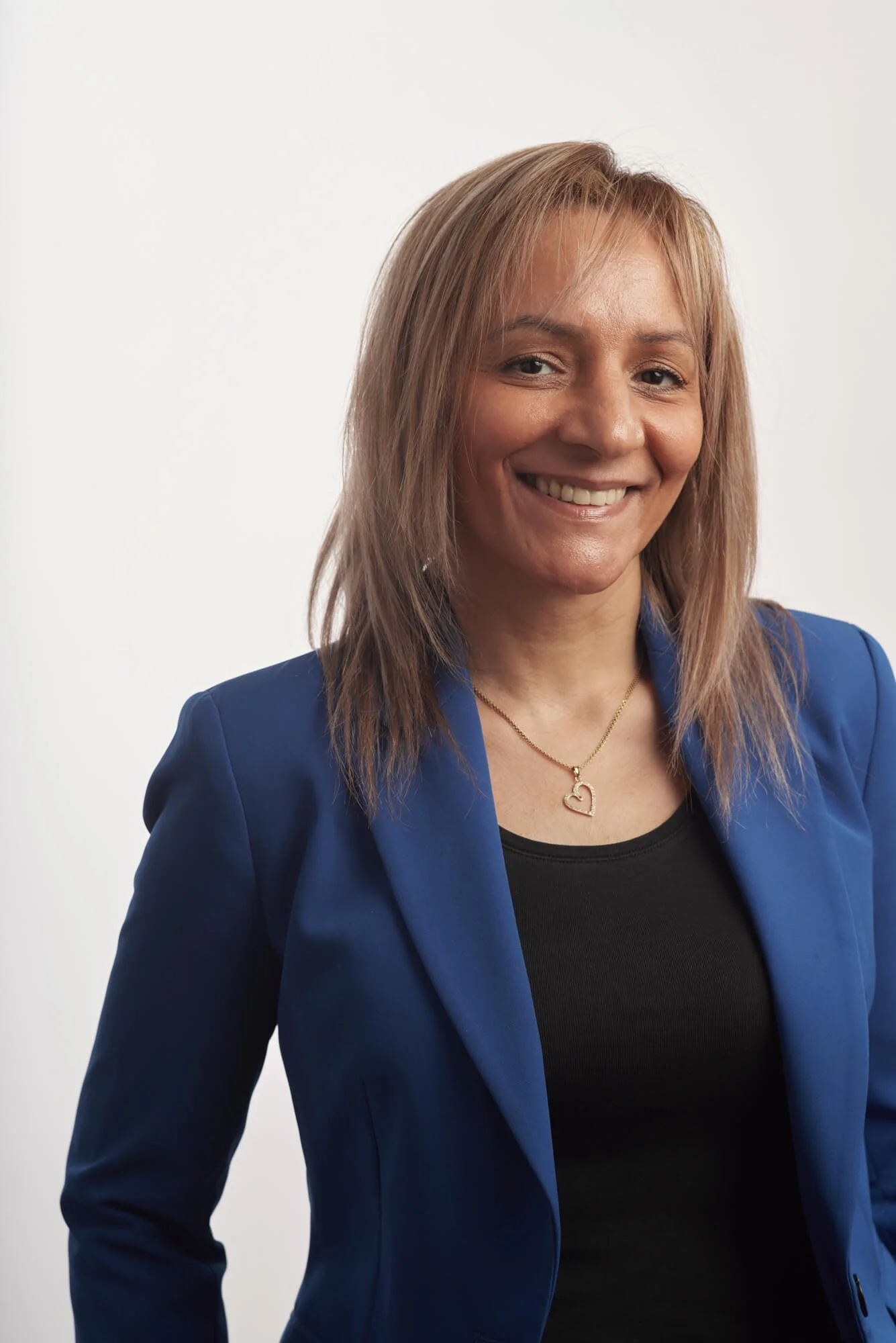 Une femme souriante portant un blazer bleu et un t-shirt noir, avec un collier en forme de cœur.