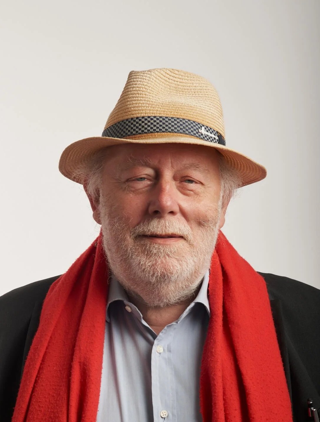 Un homme âgé souriant, portant un chapeau de paille, une écharpe rouge, une chemise blanche et une veste noire, contre un fond blanc.