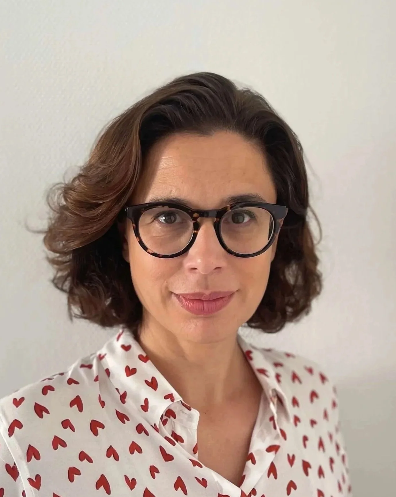 Femme portant des lunettes rondes, avec des cheveux bruns ondulés, vêtue d'une chemise blanche à motifs de cœurs rouges, souriant, contre un mur blanc.