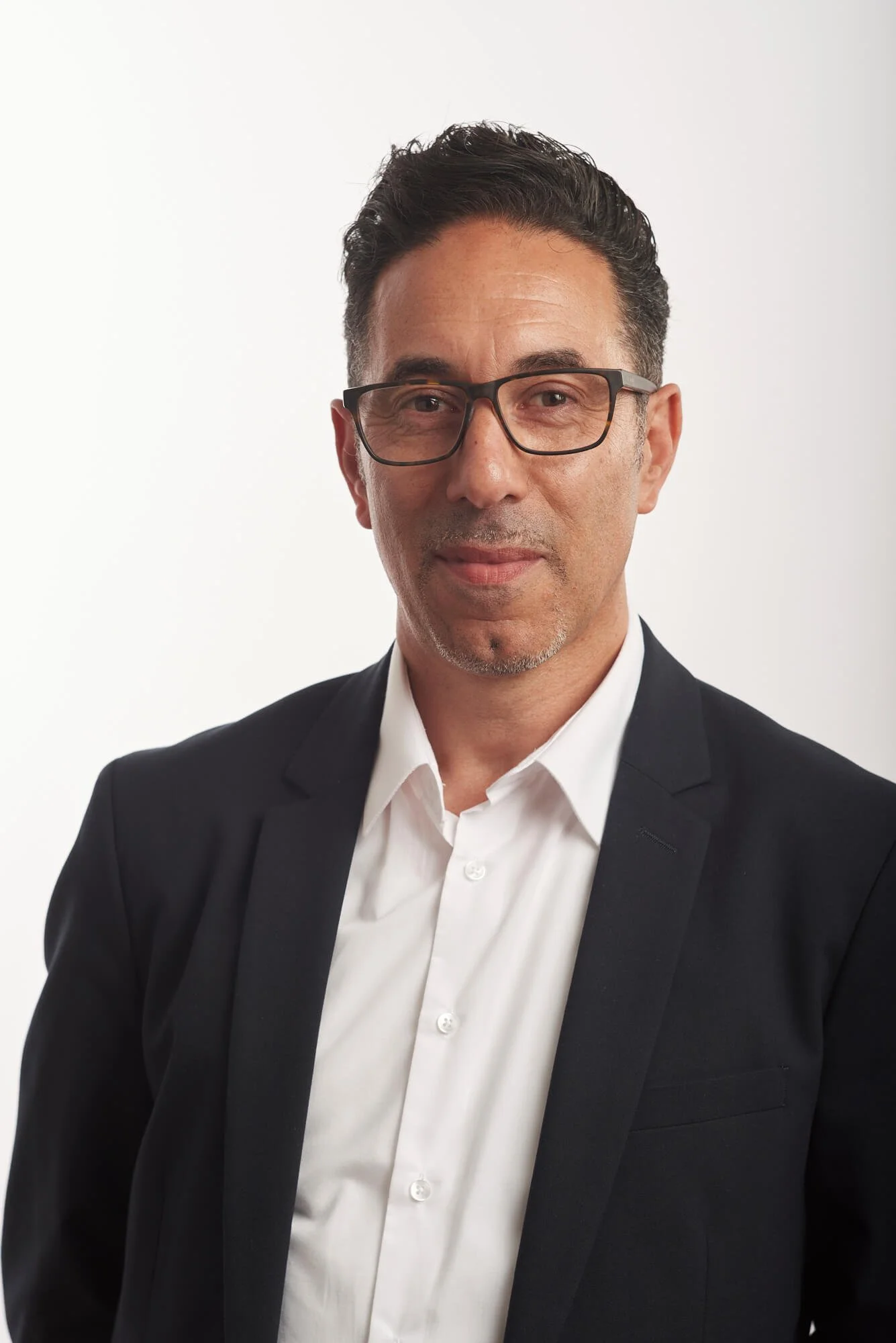 Homme d'affaires portant des lunettes, vêtu d'une chemise blanche et d'un costume noir, en pose formelle contre un fond blanc.
