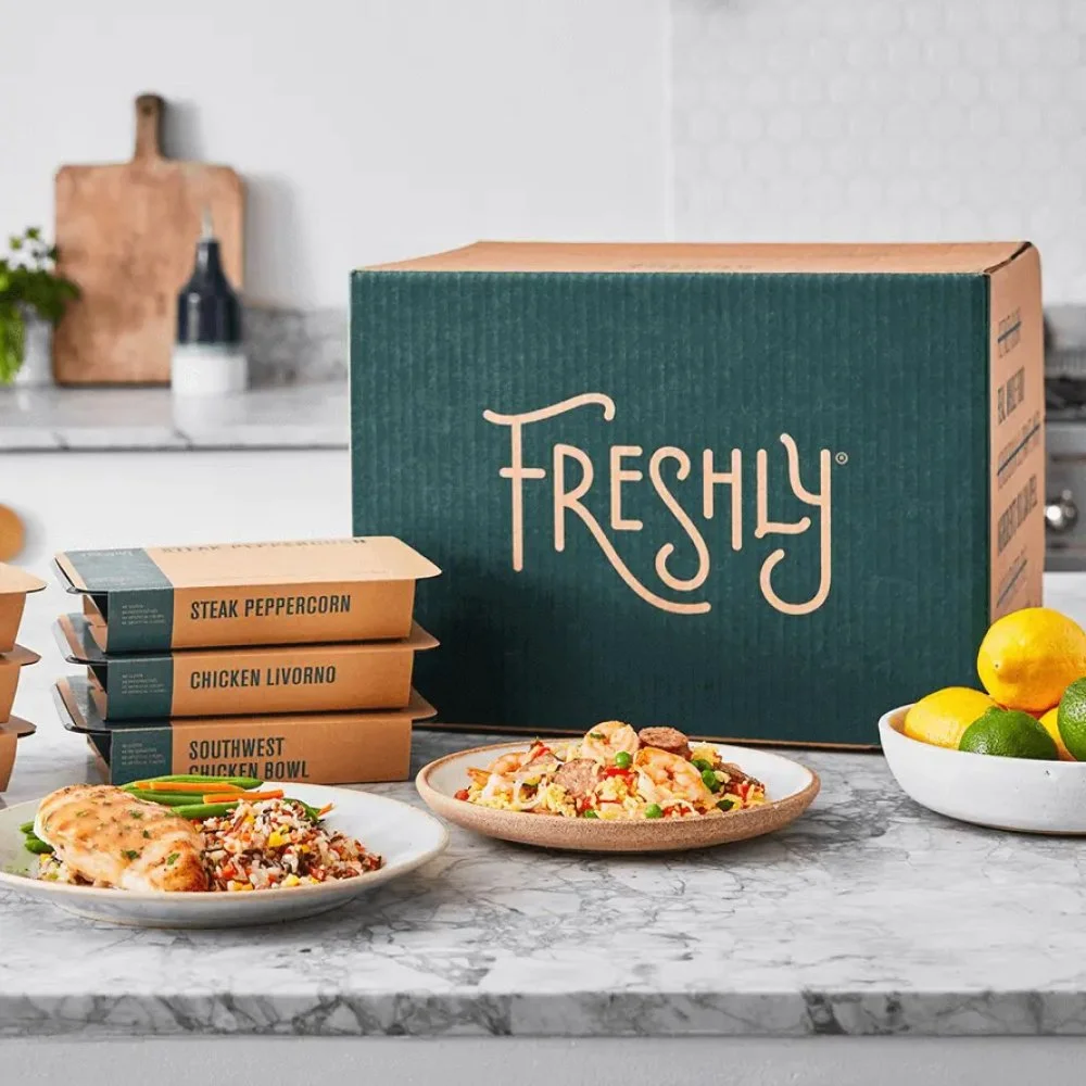 A box labeled 'Freshly' on a kitchen counter with three meal containers labeled 'Steak Peppercorn,' 'Chicken Livorno,' and 'Southwest Chicken Bowl,' along with plates of cooked food, a bowl of lemons and limes, and a cutting board with jars and a plant in the background.