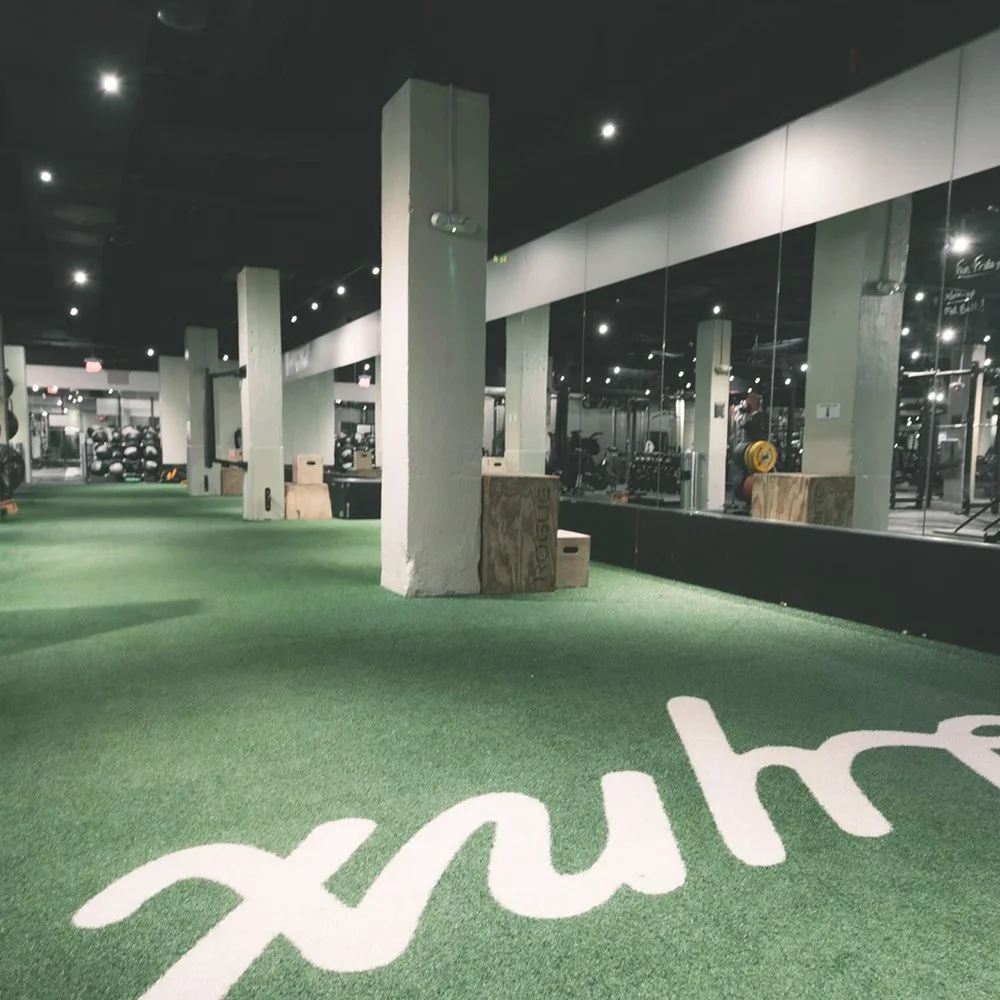 Empty gym with green artificial turf floor, gym equipment along the left wall, mirrors on the right wall, and a large word 'hustle' painted in white on the floor.
