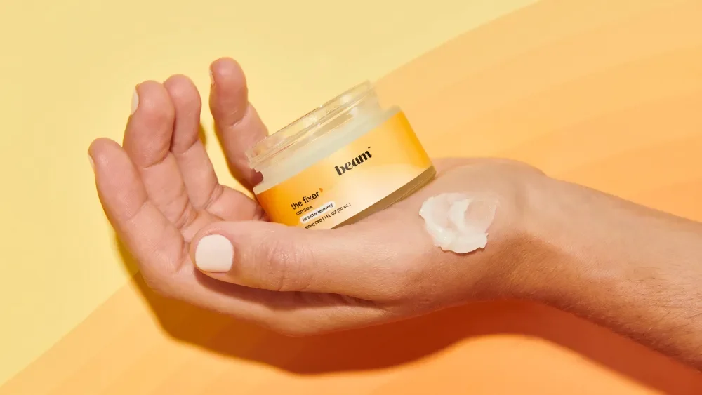A hand with a pile of cream or moisturizer on the back, holding a container labeled "beam the fixer" cream, against a yellow-orange gradient background.