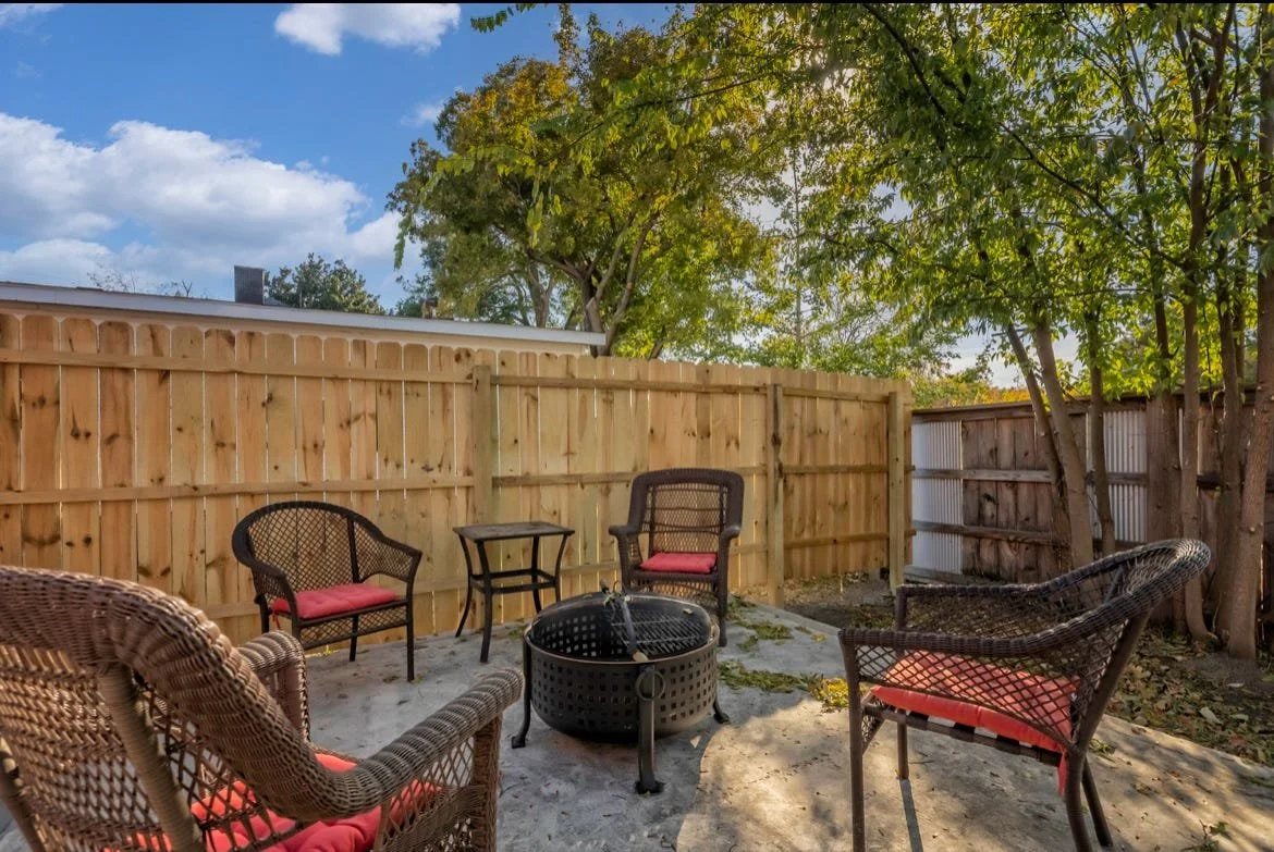 Outdoor patio with a fire pit surrounded by wicker and metal chairs with red cushions, enclosed by a wooden fence and trees, under a partly cloudy sky.
