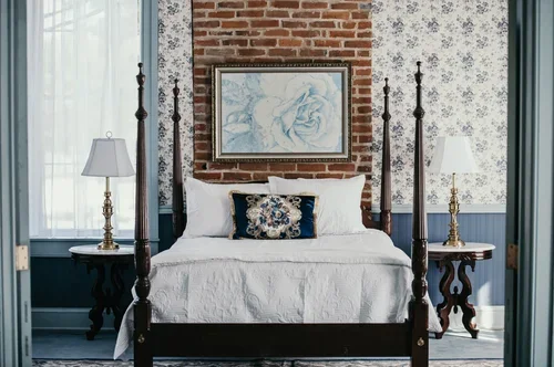 A bedroom with a four-poster bed, white bedding, decorative pillows, and wall art above the headboard, featuring brick and floral wallpapered walls.