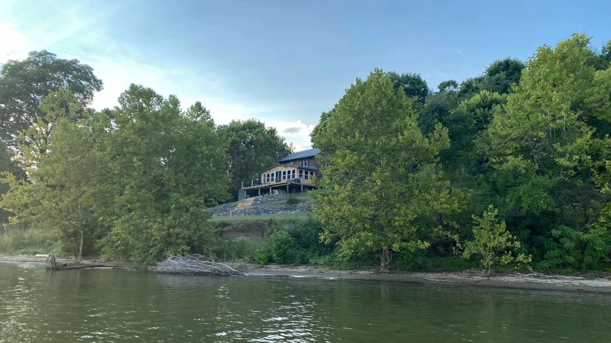 A house with a porch is situated on a hill surrounded by trees, overlooking a river or lake.