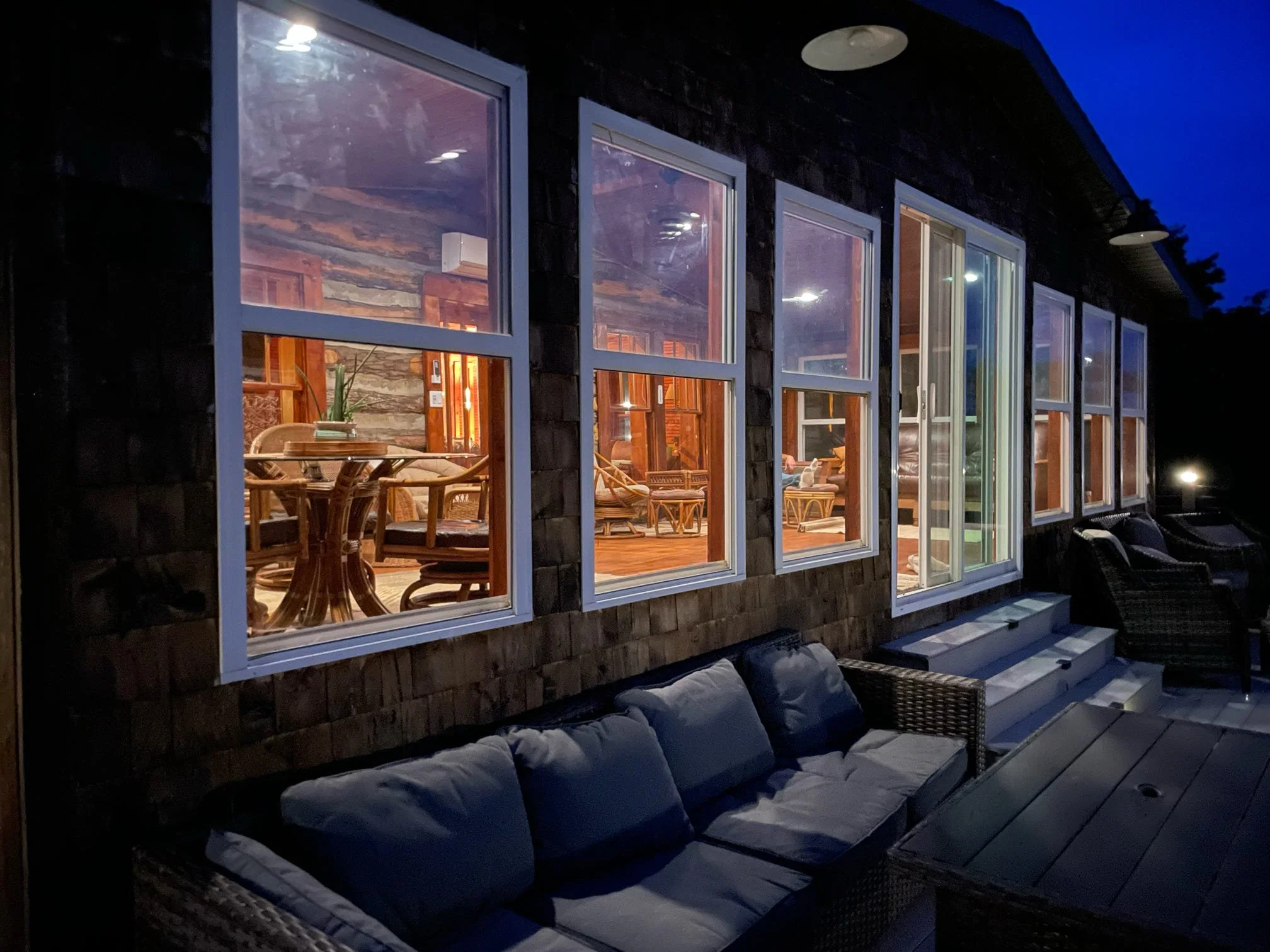 An outdoor patio at night with a cushioned bench and a large window showing a cozy indoor living space with wooden furniture and warm lighting.