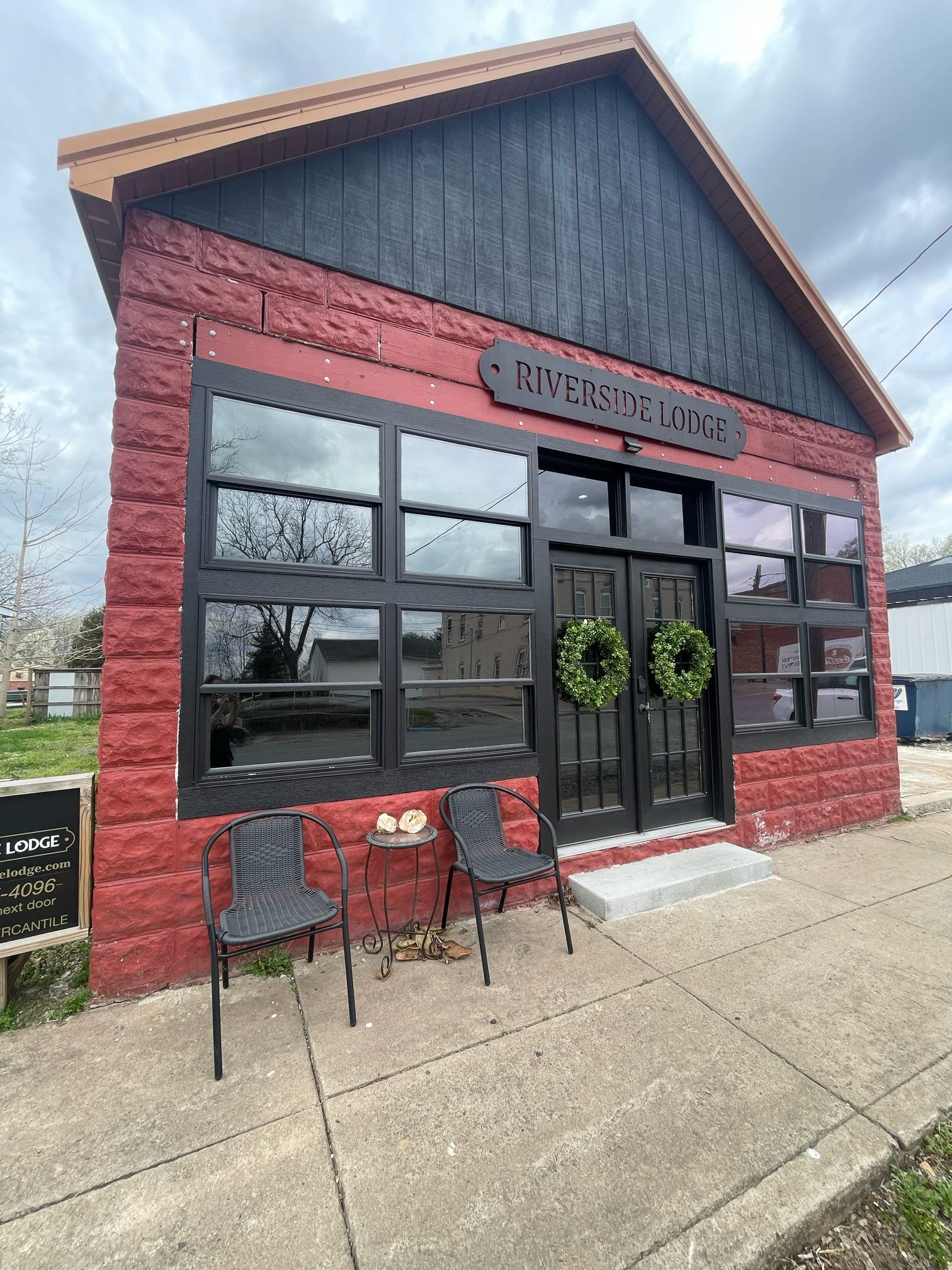 Small building with a sign that reads 'Riverside Lodge,' featuring red brick walls, black wood accents on the upper facade, a large glass garage-style door, a small patio with two chairs, and a metal roof, surrounded by trees and neighboring buildings.