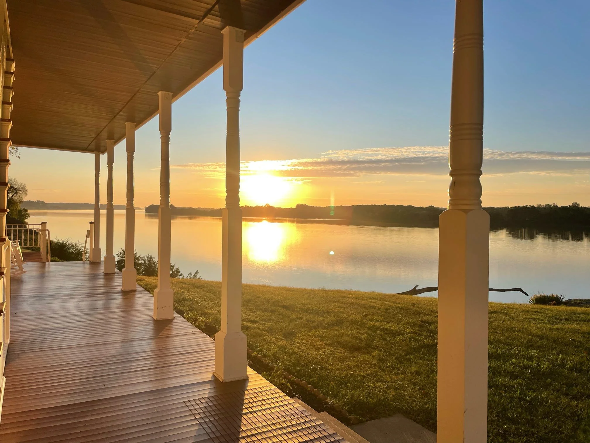 A view from a porch overlooking a body of water with a sunset in the distance. The porch has white columns and a wooden floor, with a grassy area beside it.