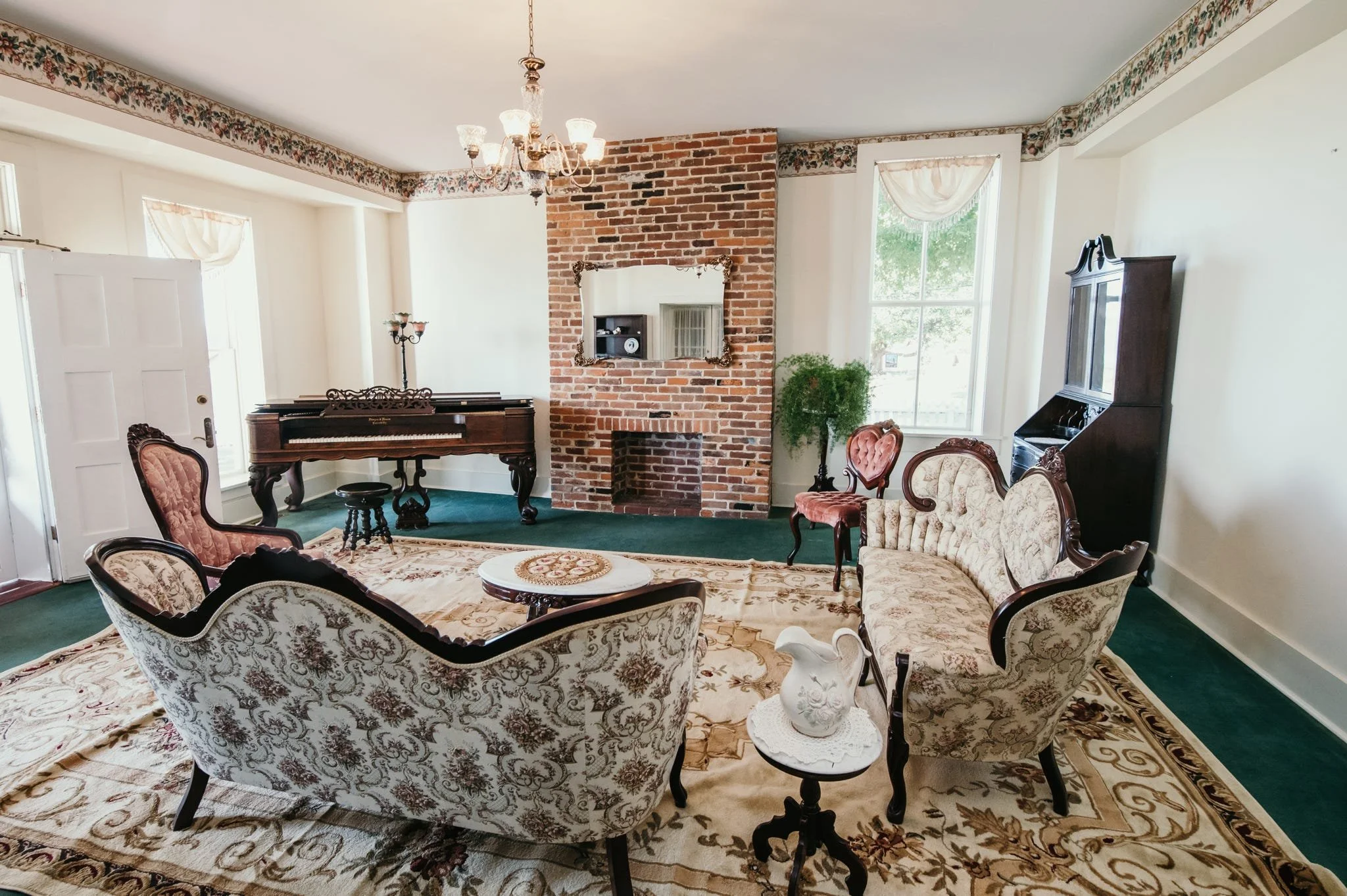 A living room with vintage furniture, a brick fireplace, large windows with curtains, and a chandelier hanging from the ceiling.