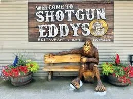 Wooden bench with a bear statue sitting next to it, surrounded by flower pots, in front of a sign for Shotgun Eddy's restaurant and bar.