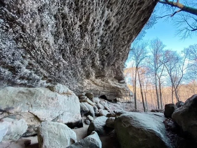 View of a large overhanging rock formation with smaller rocks and a dry creek bed underneath, with leafless trees and a clear blue sky in the background.