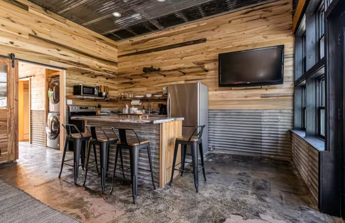 Kitchen with wooden walls, a large sink, stainless steel refrigerator, black bar stools at a marble countertop, and a wall-mounted flat-screen TV.