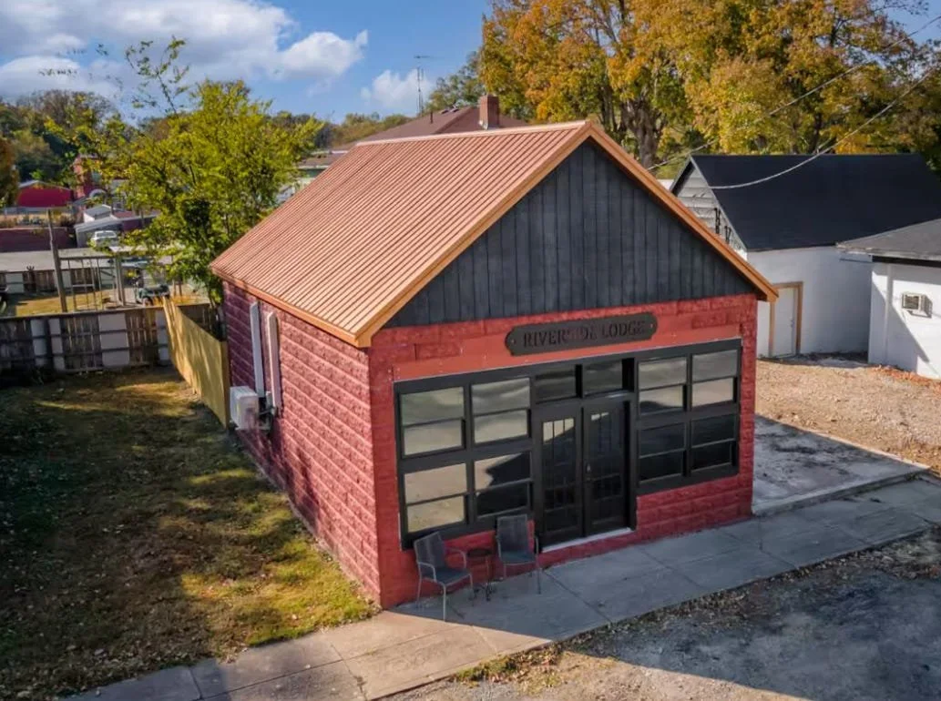A small commercial building with a brick exterior, sign reading 'Riverside Lodge,' large shop windows, a metal roof, and two chairs outside on a concrete sidewalk, with trees and other buildings in the background.