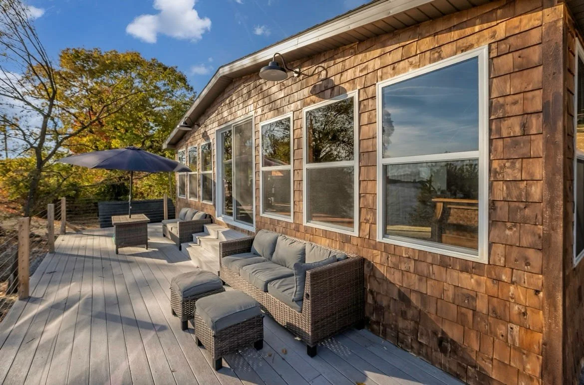 Outdoor wooden deck with wicker furniture, including a sofa, ottomans, and chairs, with a large umbrella, attached to a house with wood shingle siding and multiple windows, surrounded by trees.