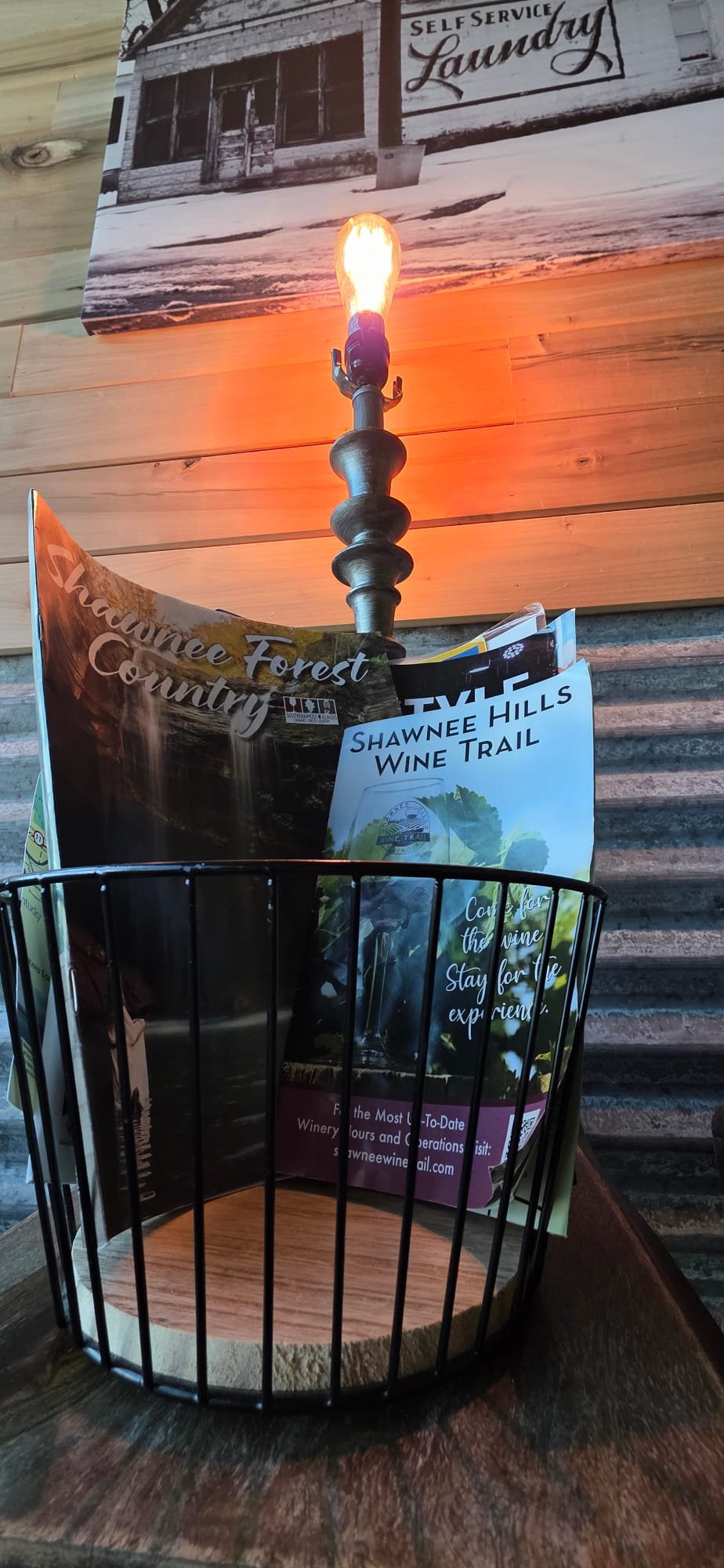 A decorative lamp turns on above a table with brochures about Shawnee Hills wine trail, set against a wooden wall.