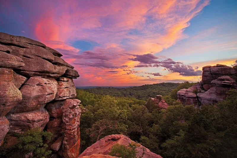 Sunset over a landscape with rock formations and green trees.