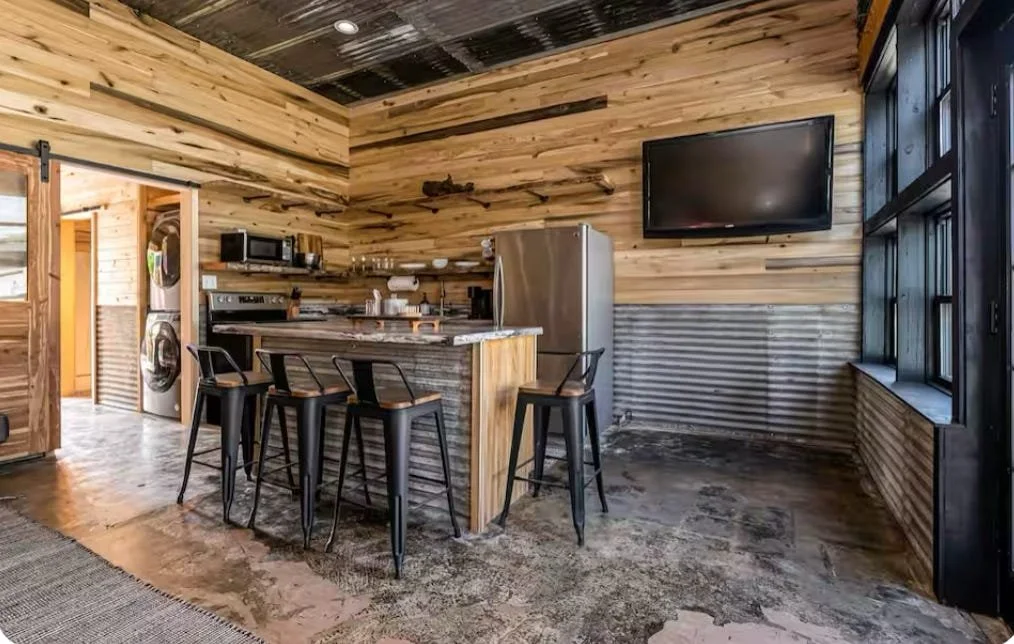 Rustic kitchen with wood-paneled walls, a marble-topped island with black chairs, stainless steel refrigerator, and a wall-mounted TV, featuring large windows.
