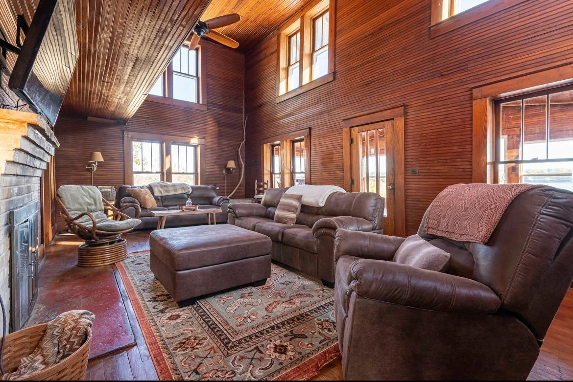 Living room with wooden walls, high ceiling, large windows, leather furniture, a patterned rug, fireplace, ceiling fan, and natural light.