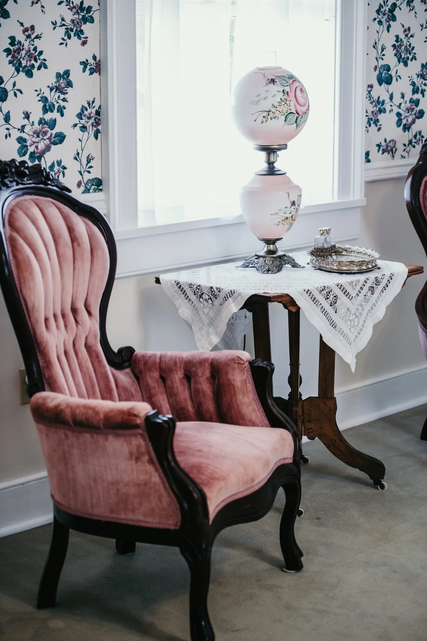 A vintage pink velvet armchair with dark wooden legs and carved frame, positioned near a small wooden table with a lace tablecloth, a decorative lamp with floral design, and a glass perfume bottle on top. The room has floral wallpaper and white windo